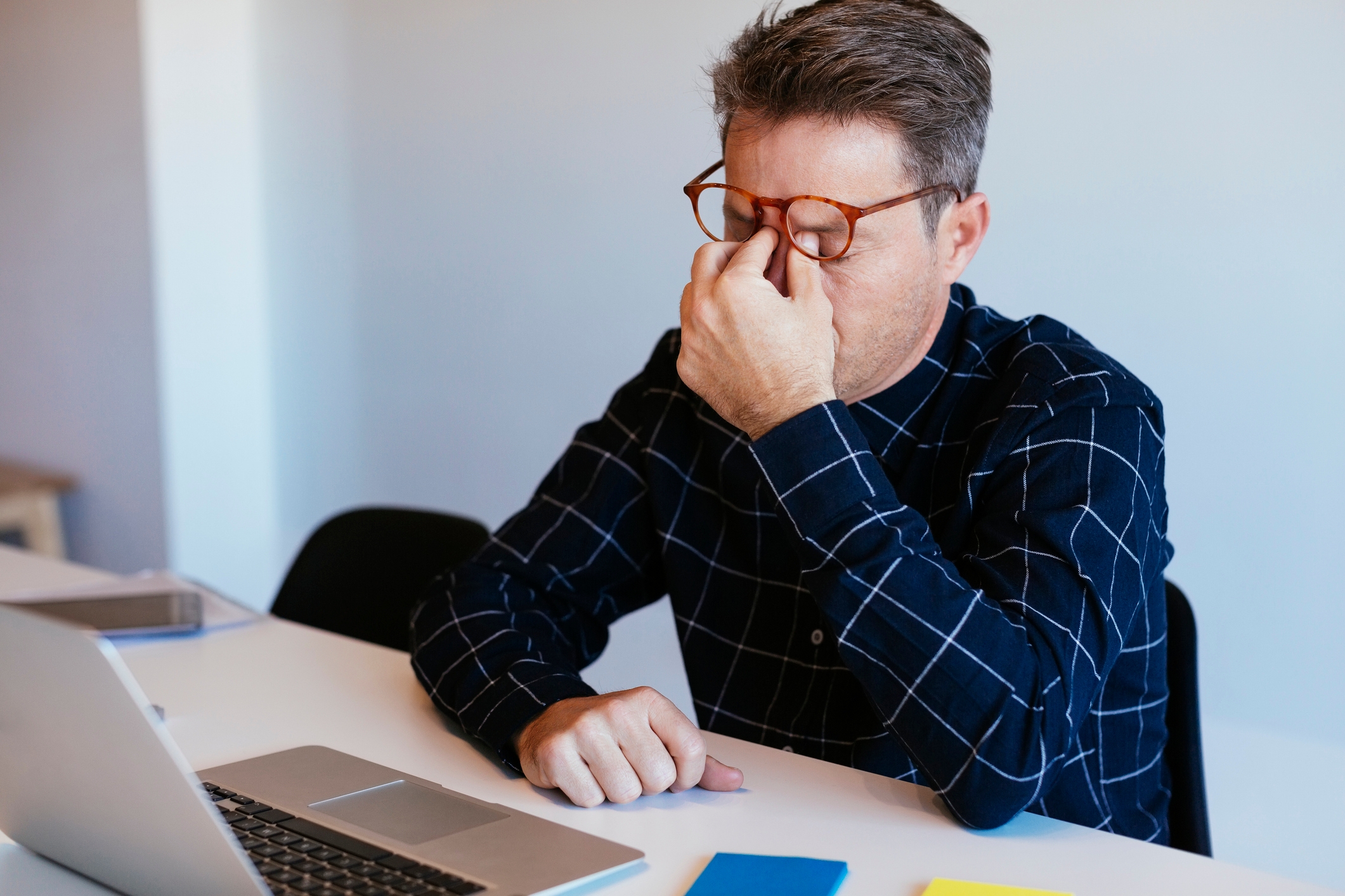 Person in a checkered shirt sits at a desk, rubbing their eyes while looking at a laptop, suggesting fatigue or eye strain