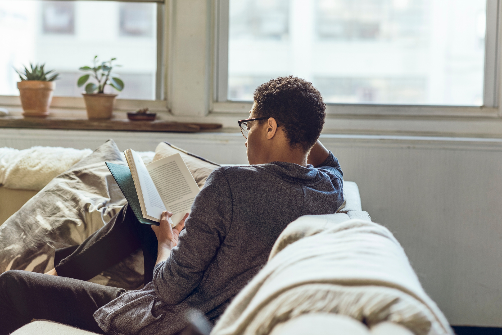 Person reading a book on a cozy, light-filled sofa with plants on the windowsill