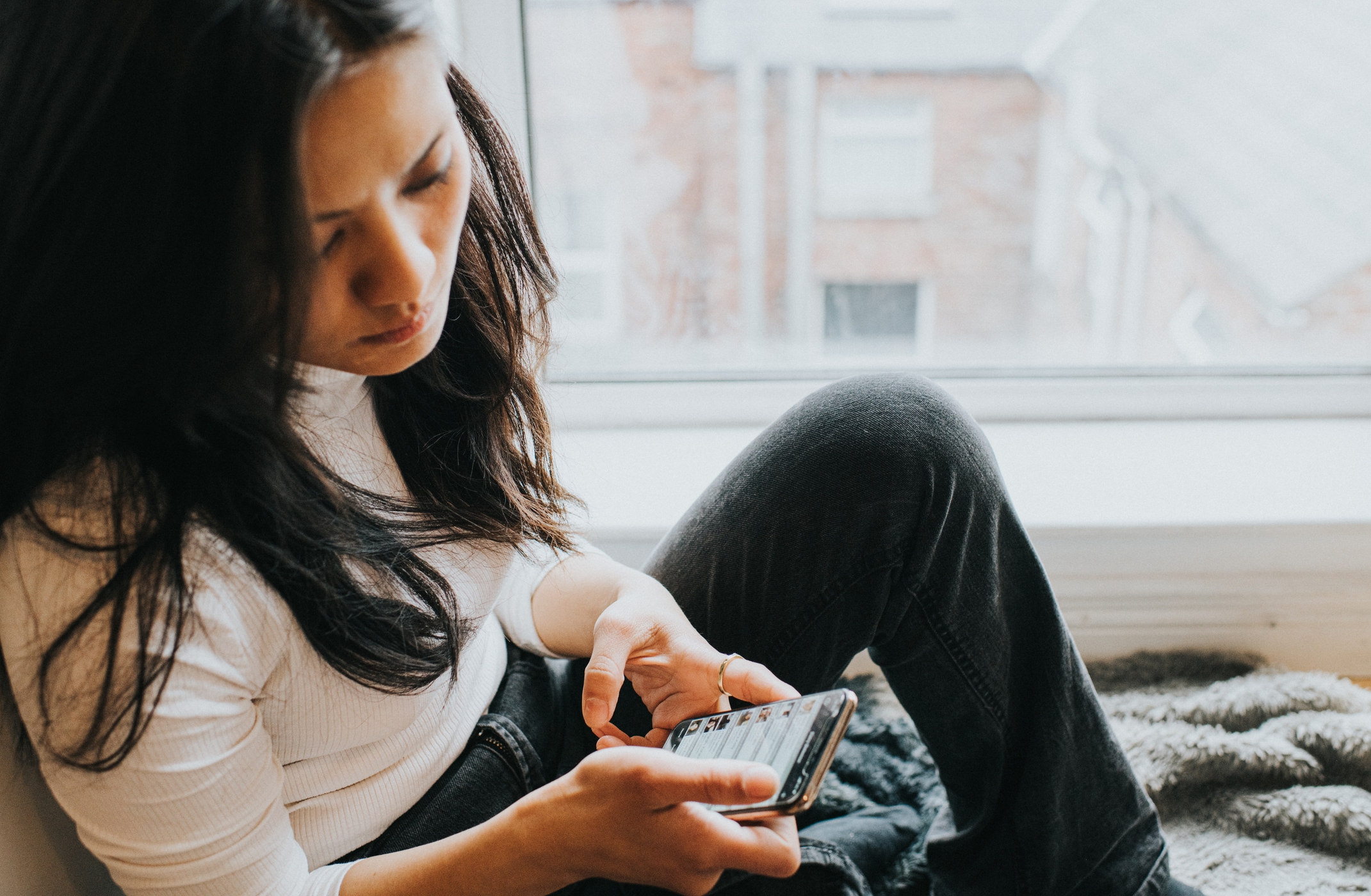 Person seated by a window, focused on their phone screen, wearing a white top and jeans. Relaxed, introspective mood