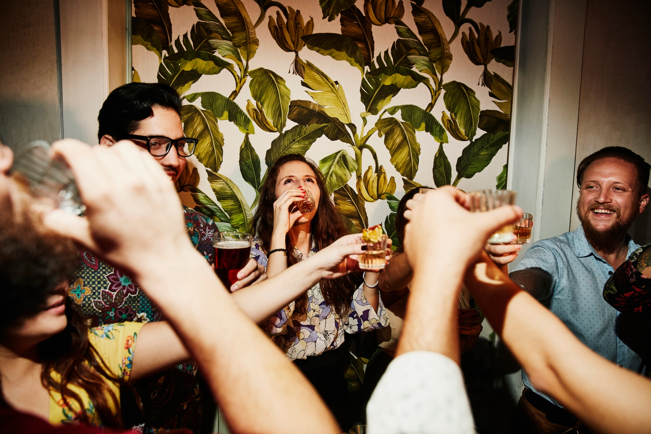 People raising glasses in a toast at a lively indoor party, surrounded by friends and a tropical leaf-patterned wall
