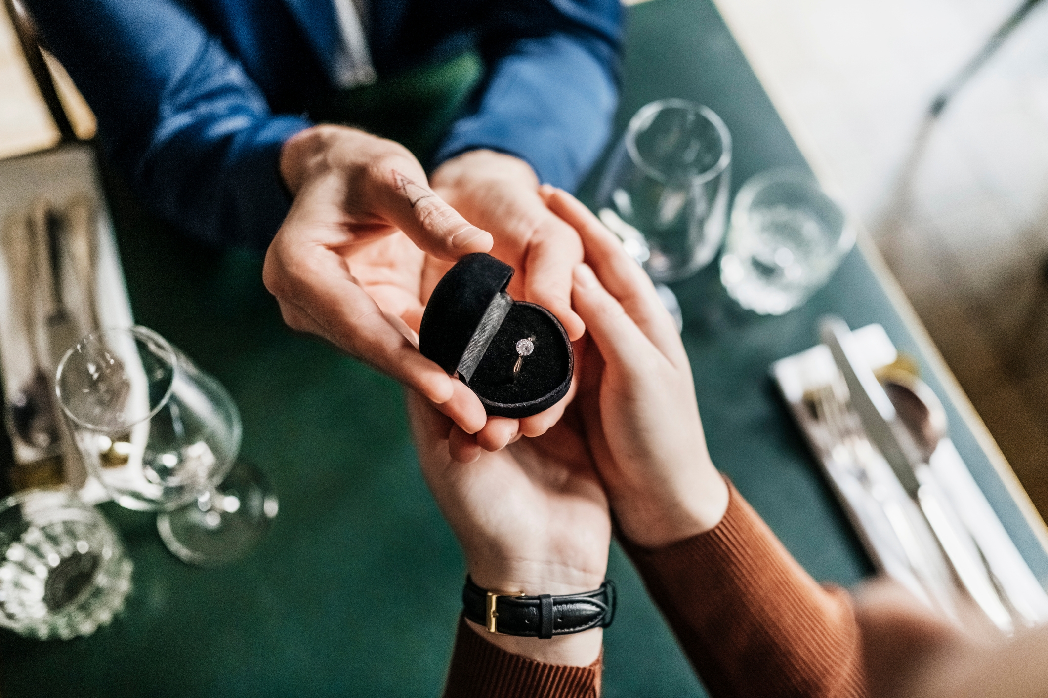 A person holds an open ring box presenting a diamond engagement ring to another person across a dining table, suggesting a proposal