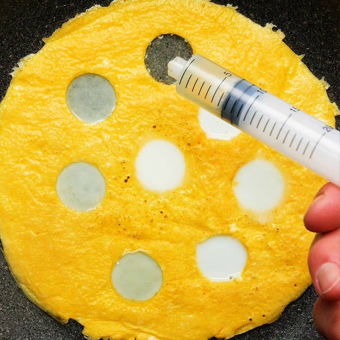 フライパンで調理中の卵液に、注射器で円形の穴を開ける手元。