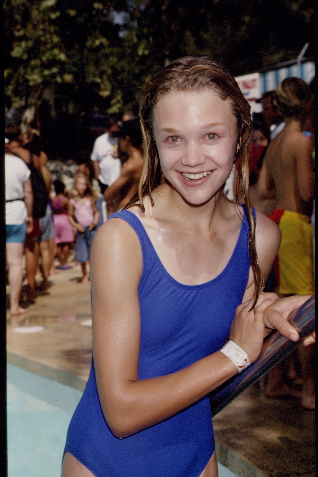Person smiling in a pool area, wearing a one-piece swimsuit. Others are in the background, suggesting a casual, outdoor water event