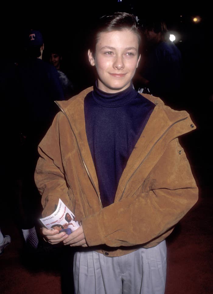 Young person on red carpet, wearing a turtleneck and jacket, holding a pamphlet