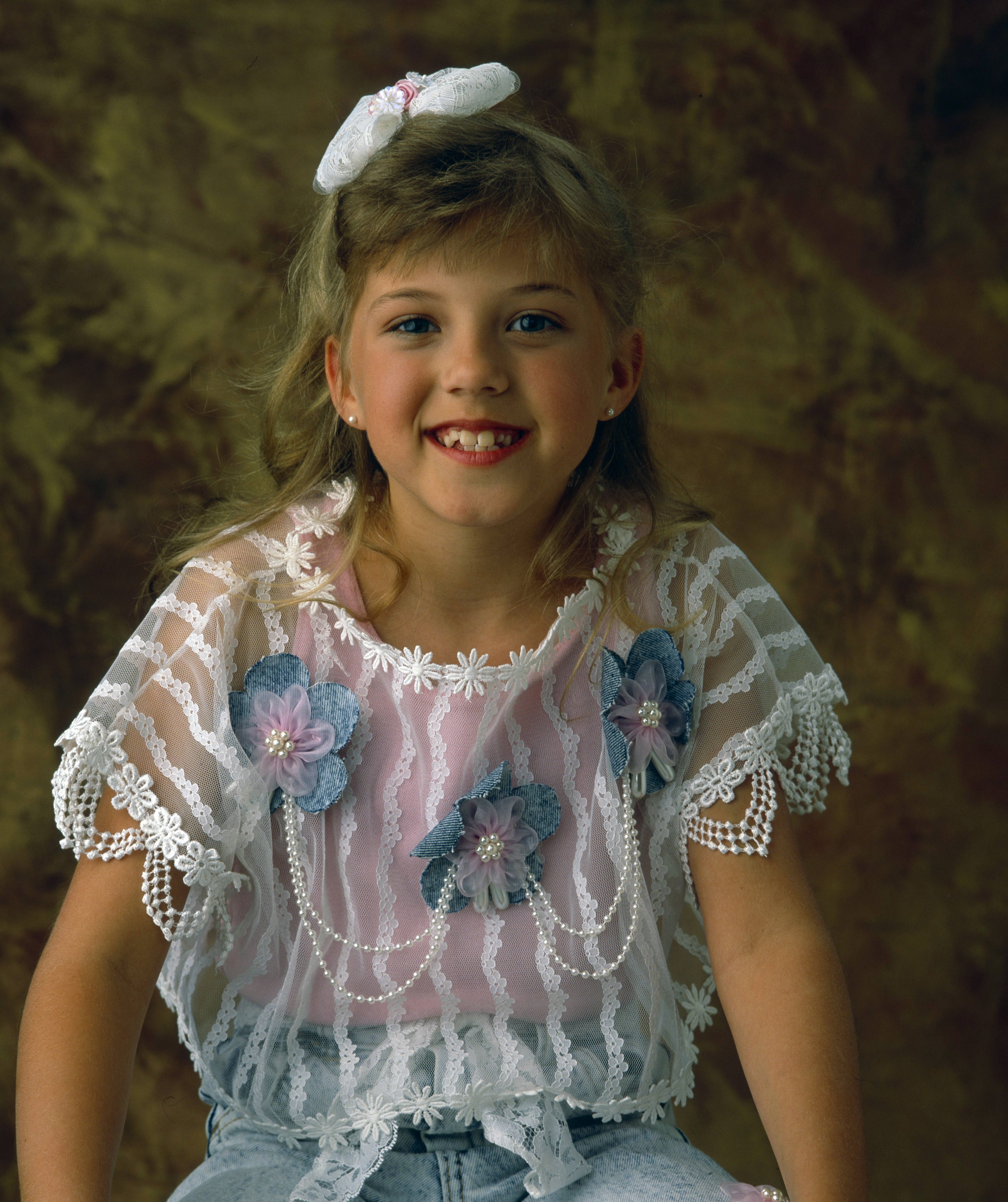 Young girl smiling, wearing a lace top with floral accents and a hair bow, sitting indoors