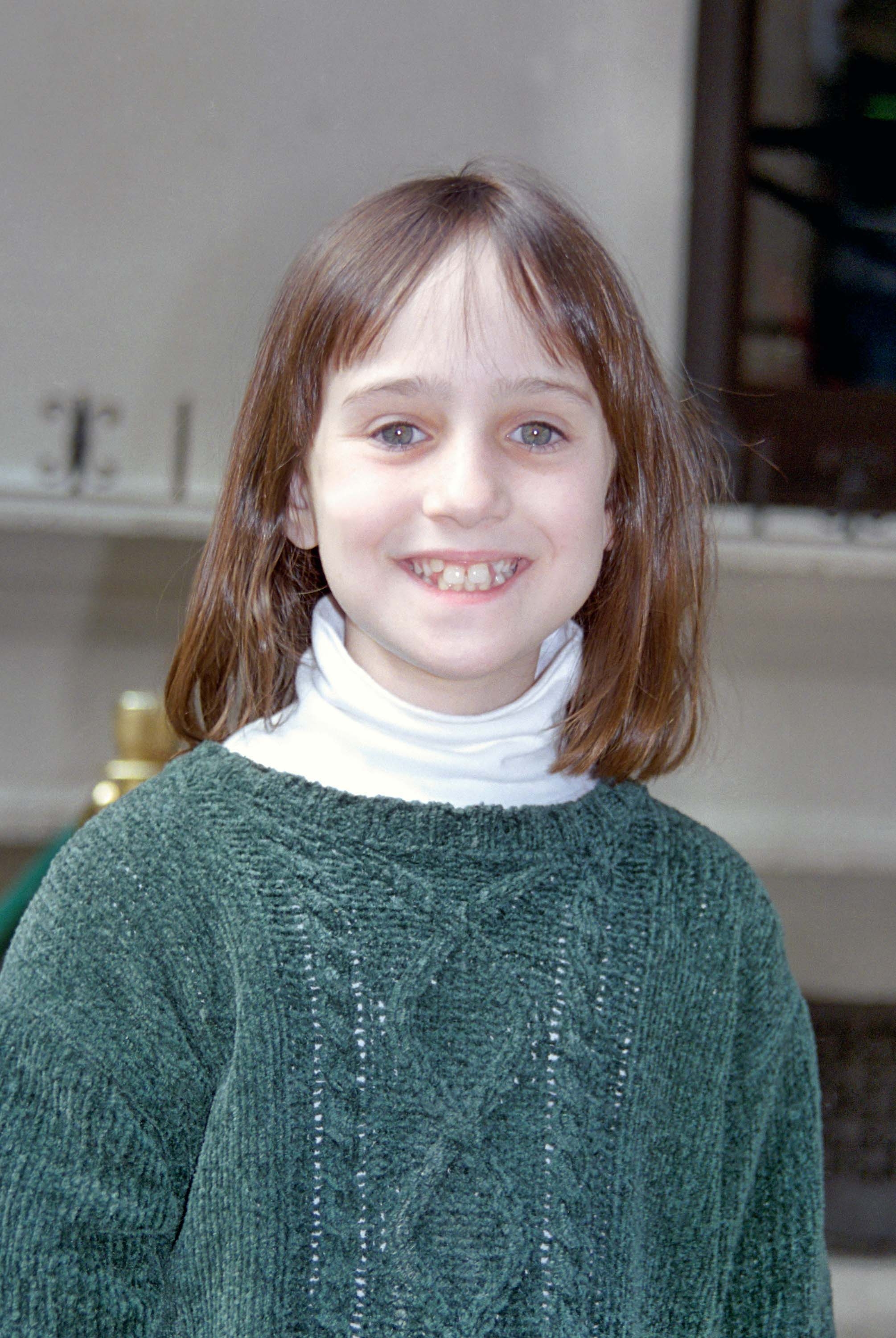 A young girl with shoulder-length hair smiles, wearing a thick ribbed sweater over a turtleneck