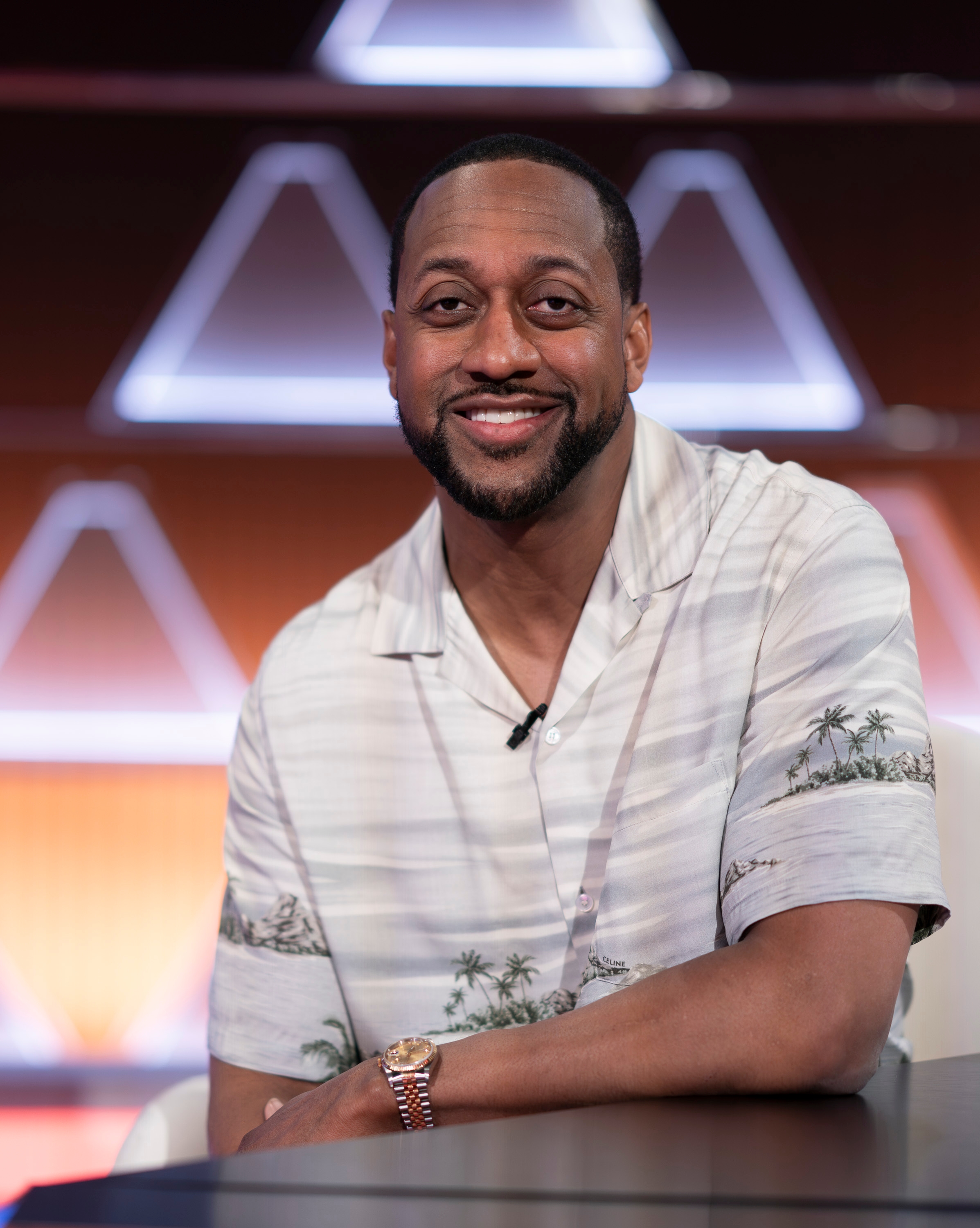 Person wearing a casual, buttoned shirt with palm tree design, seated and smiling in front of a geometric background