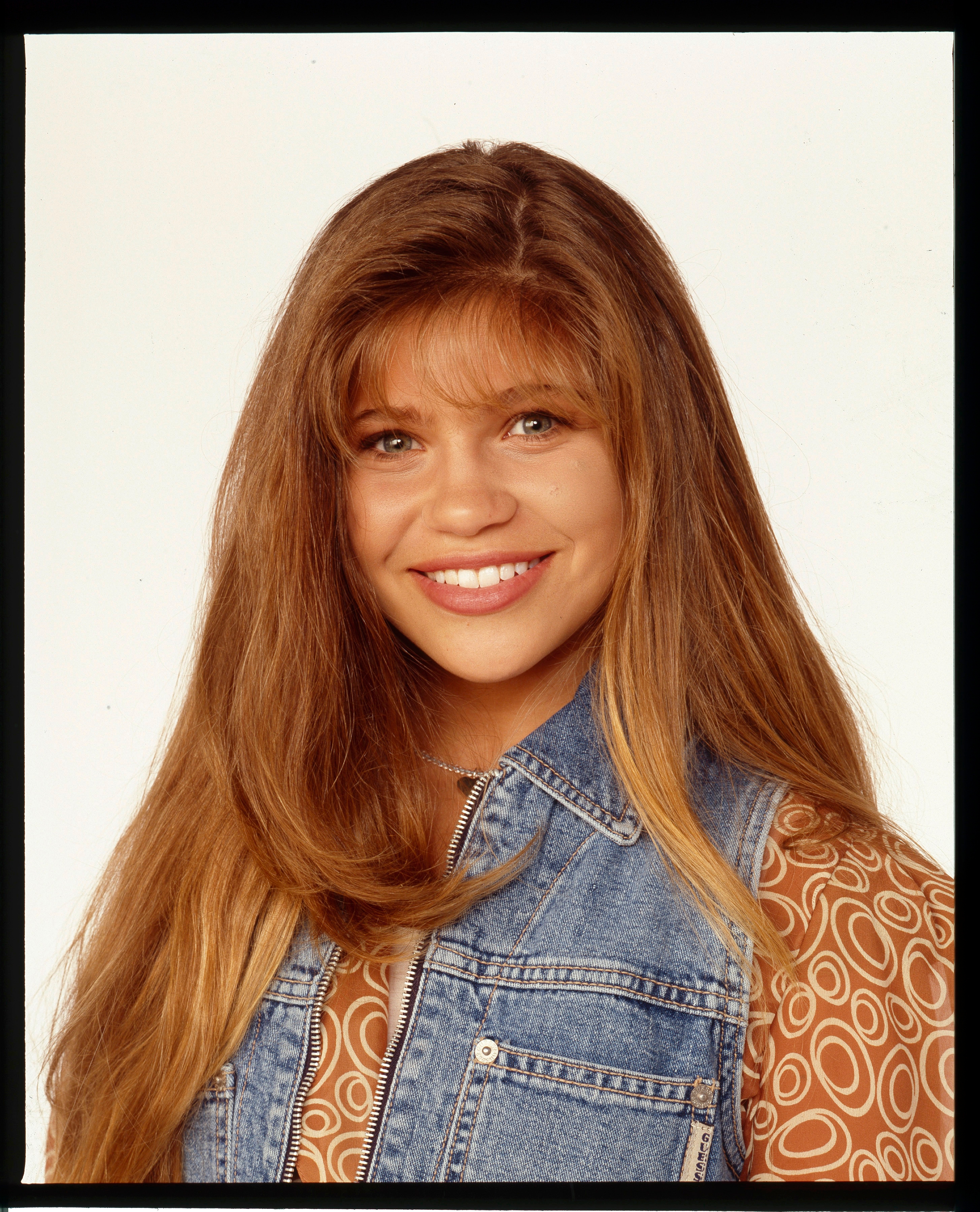 Smiling person with long hair wearing a denim vest over a patterned top