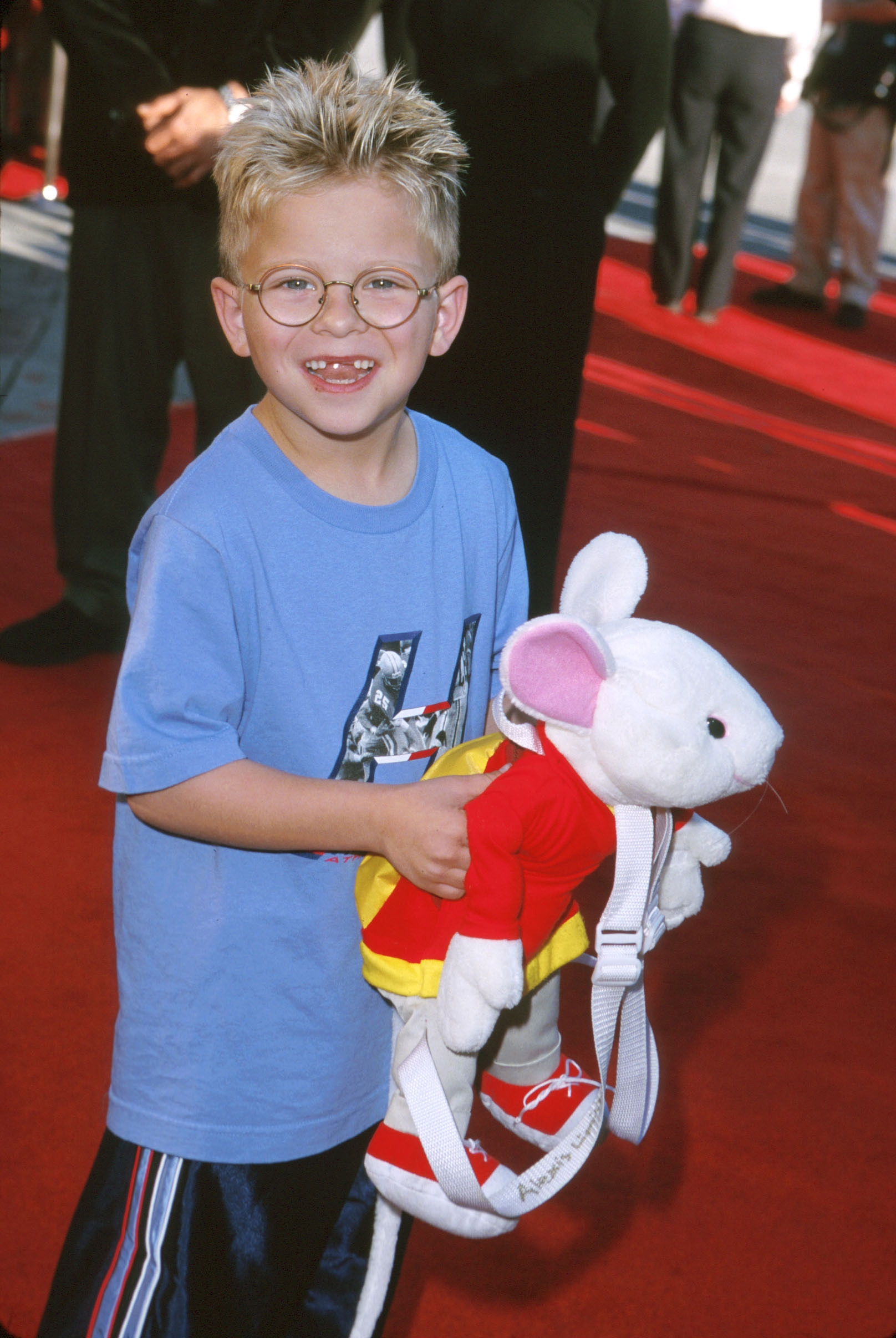 A young child on a red carpet holds a plush mouse backpack. The child smiles, wearing a casual T-shirt and glasses