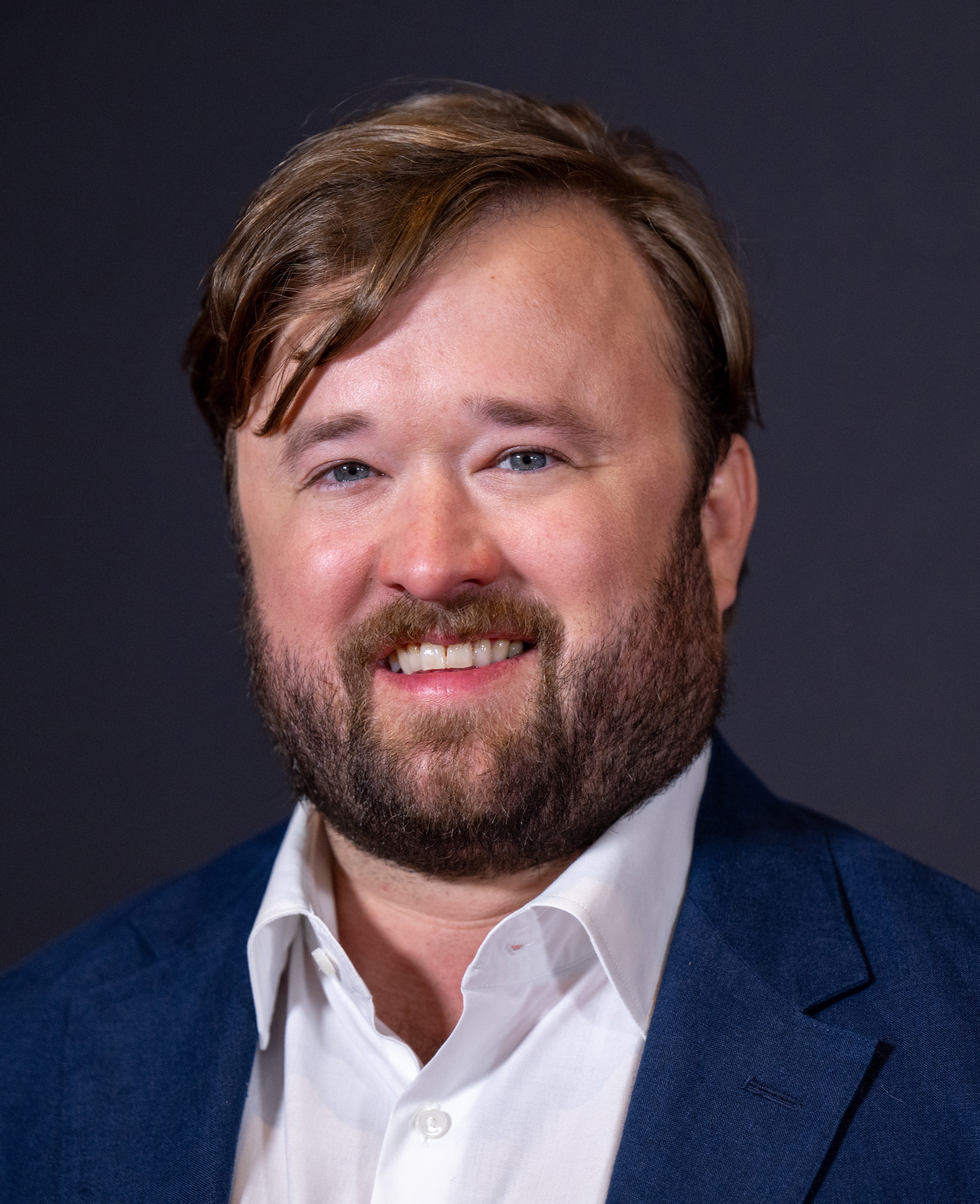 Man with a beard and styled hair in a blazer and white shirt, smiling at the camera
