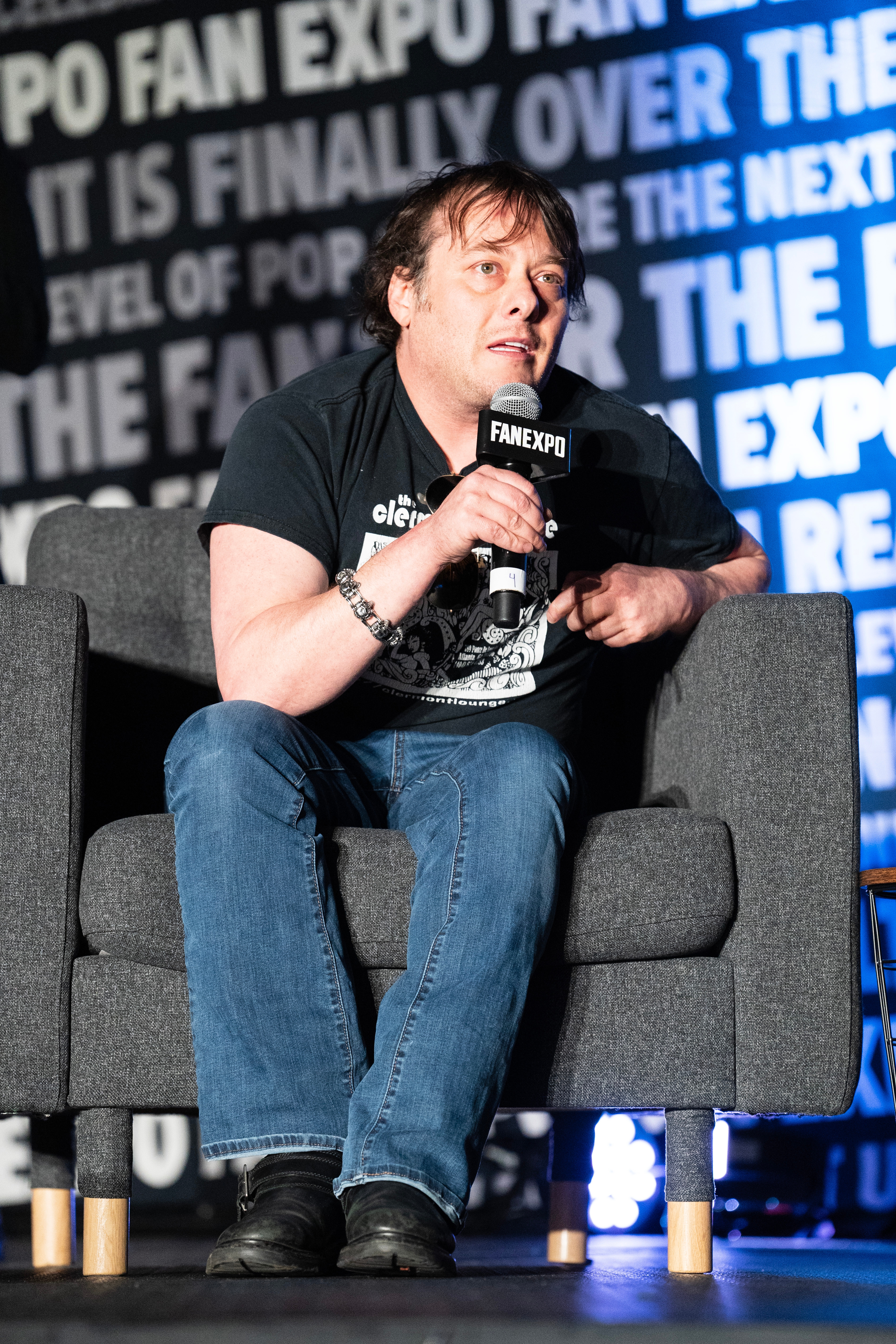 Person seated at a fan expo panel holding a microphone, wearing a printed t-shirt and jeans, speaking to the audience