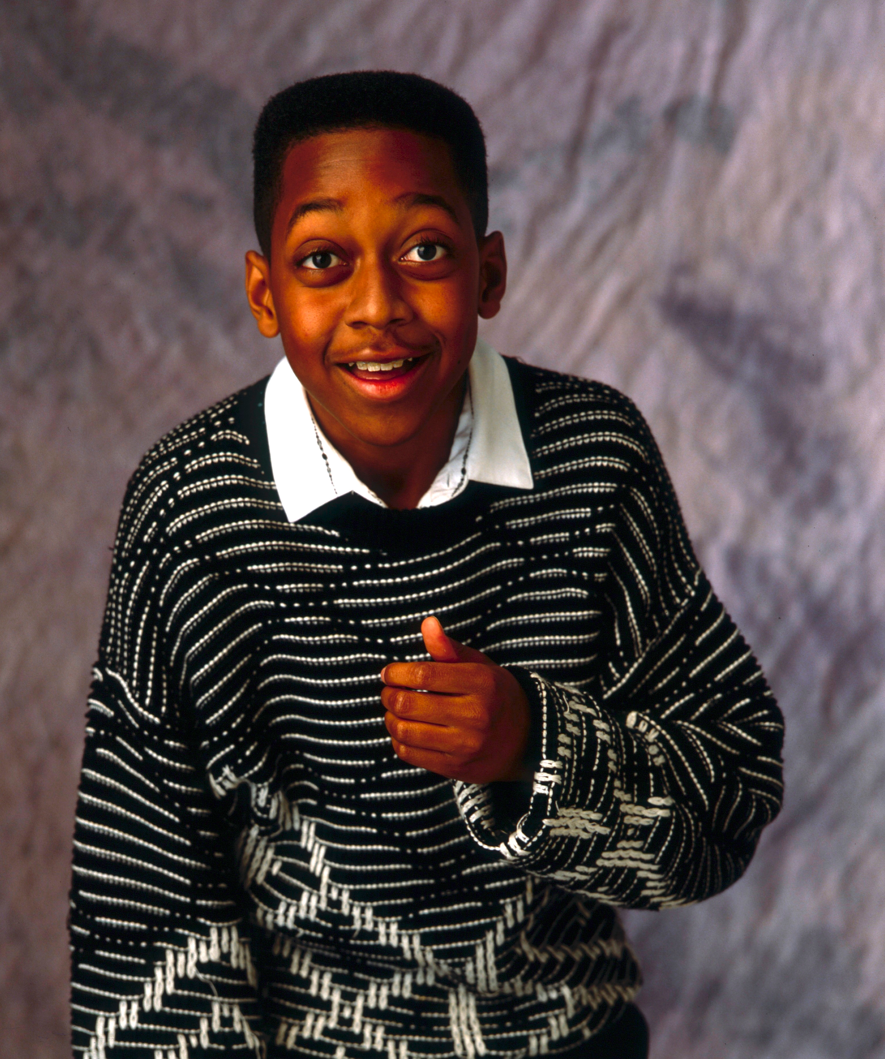 Person wearing a patterned sweater, smiling and gesturing with one hand against a textured background