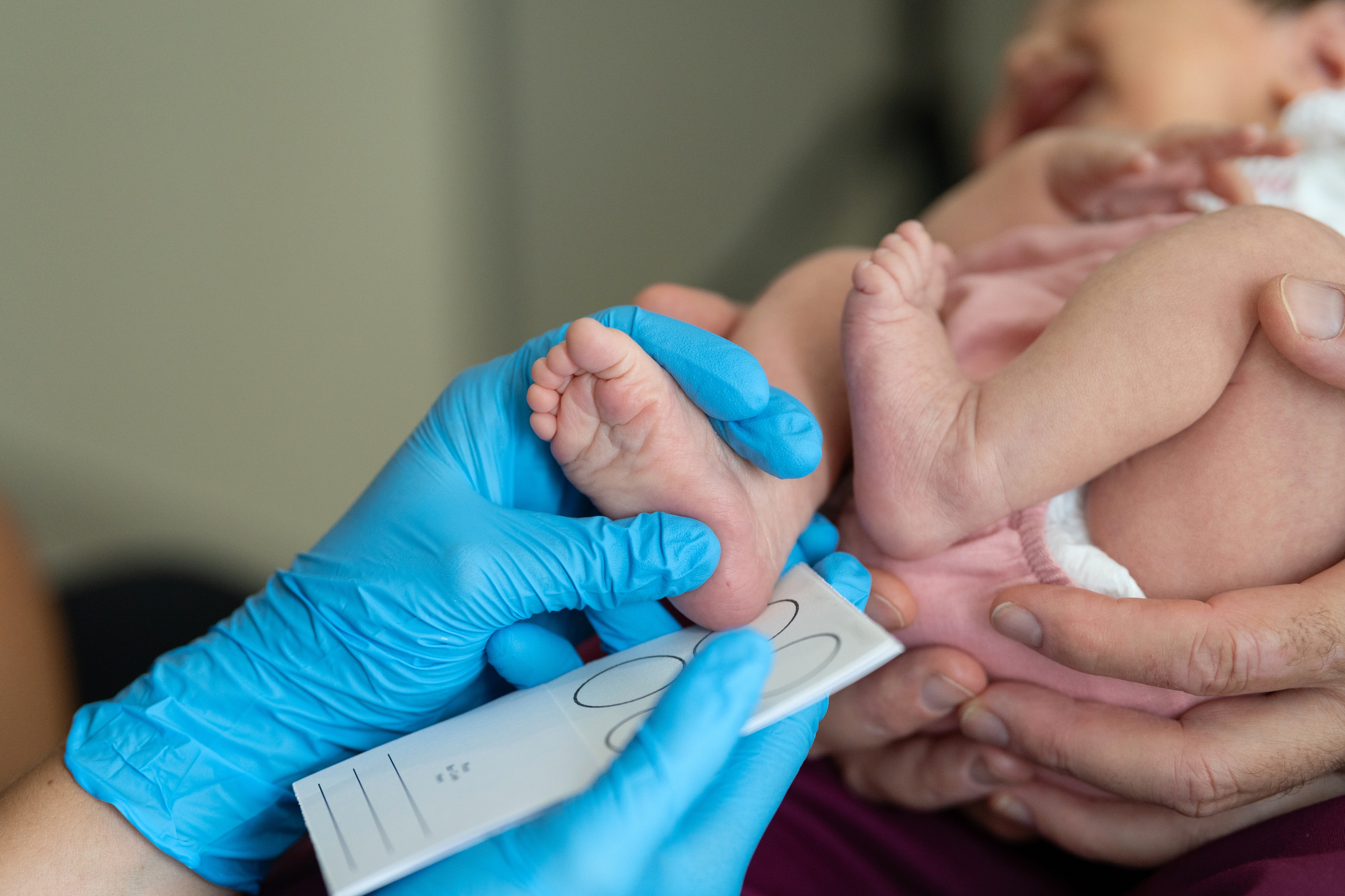 A healthcare worker in gloves is taking a newborn's footprints on a card while an adult holds the baby