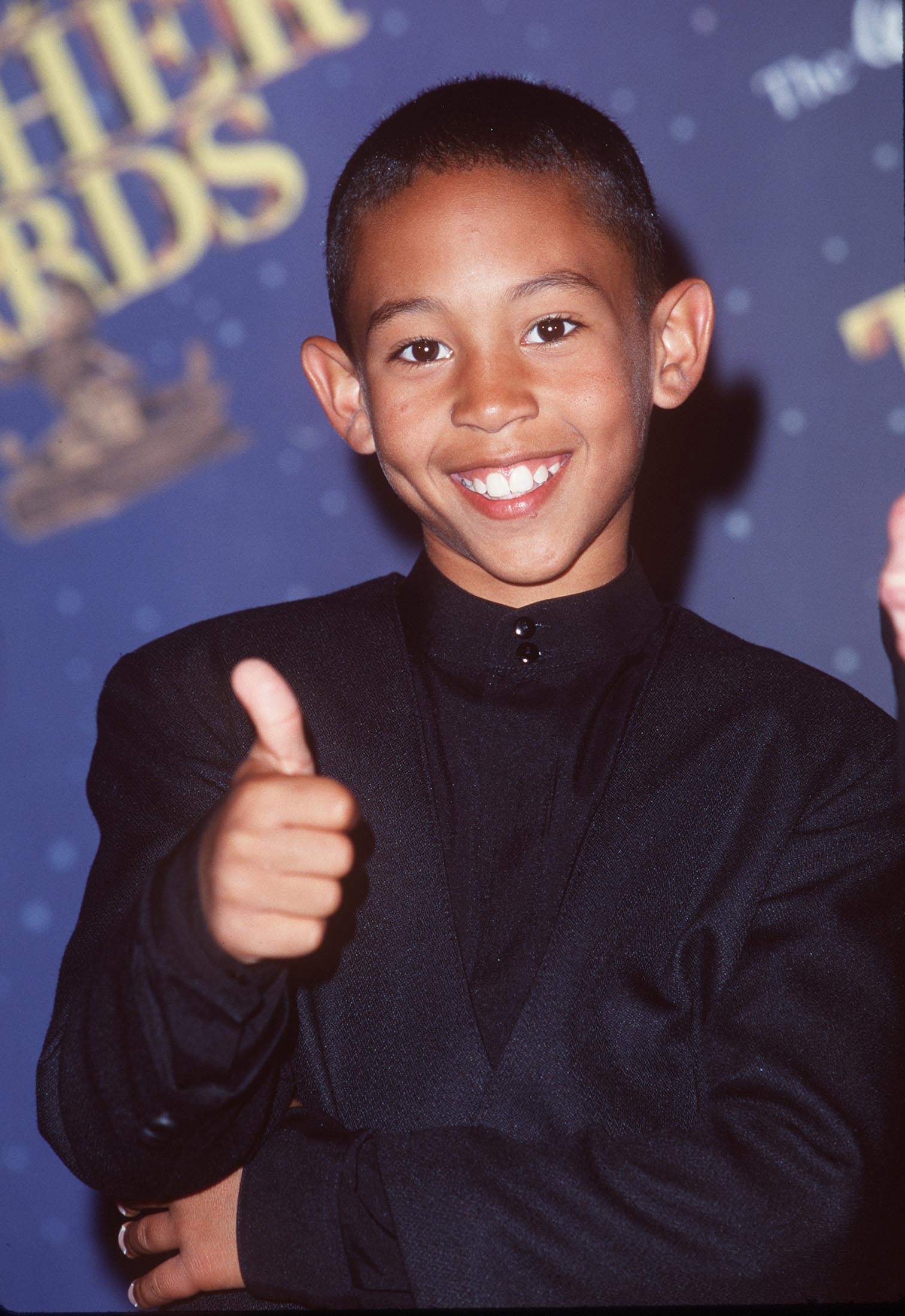 Person smiling and giving a thumbs-up on a red carpet, wearing a sleek, high-collared jacket