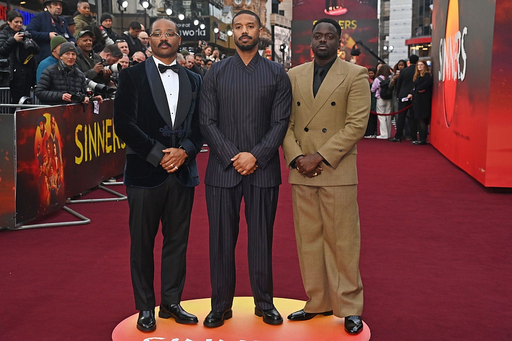 Three men in formal attire pose on a red carpet at a movie event with a crowd and a poster in the background