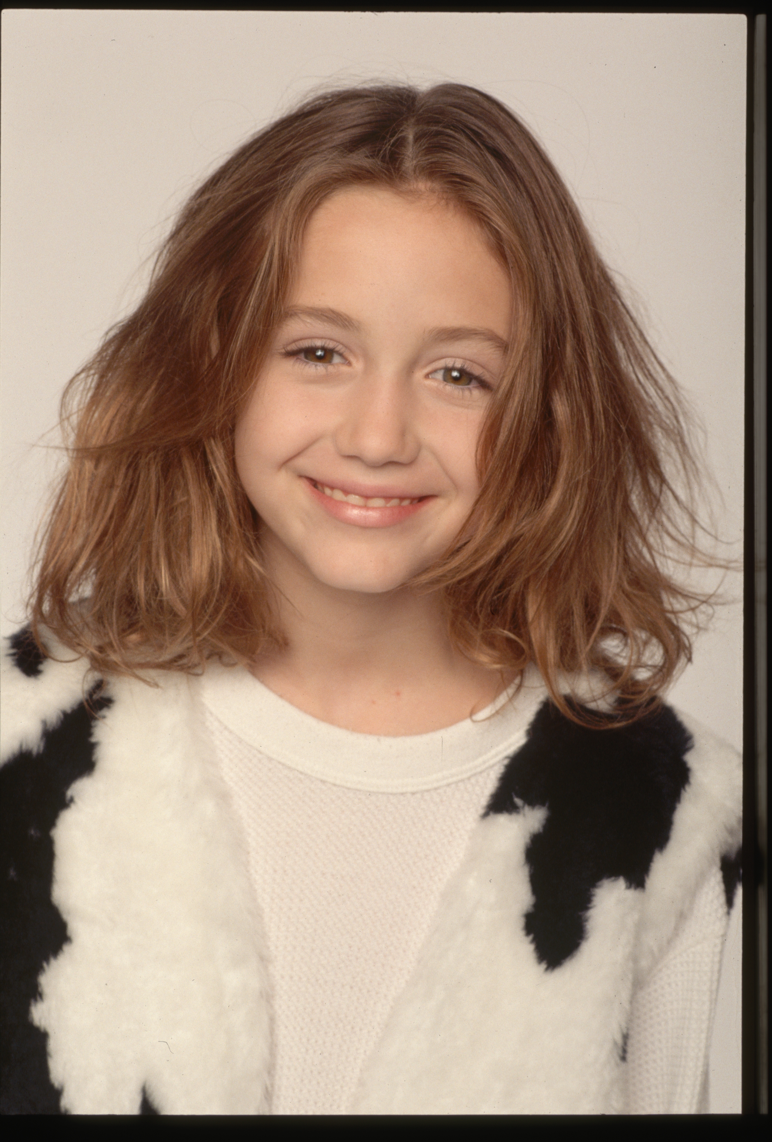 Person smiling with shoulder-length hair, wearing a patterned vest over a shirt, looking directly at the camera