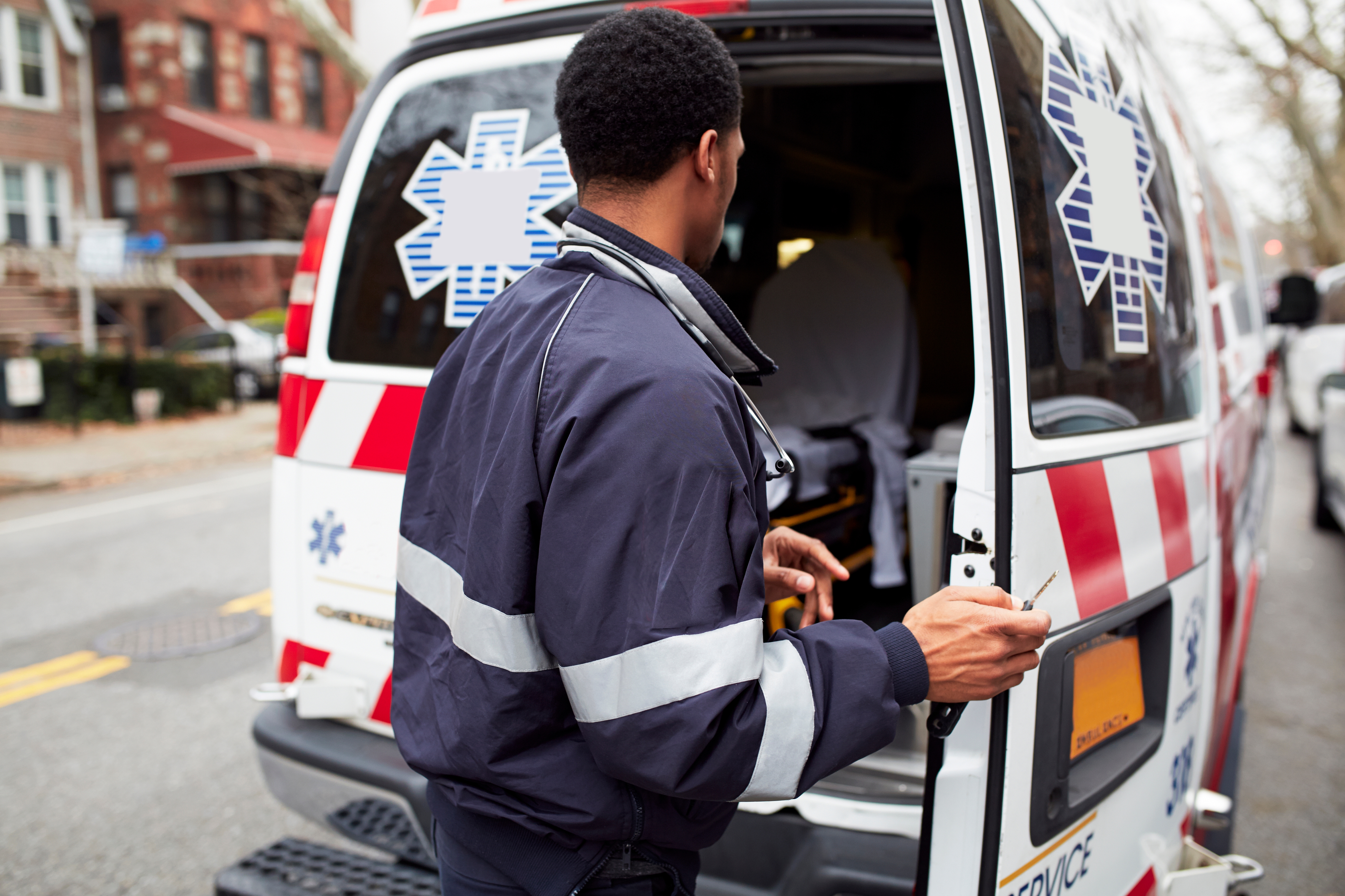 A paramedic stands at the back of an ambulance, preparing equipment on a residential street