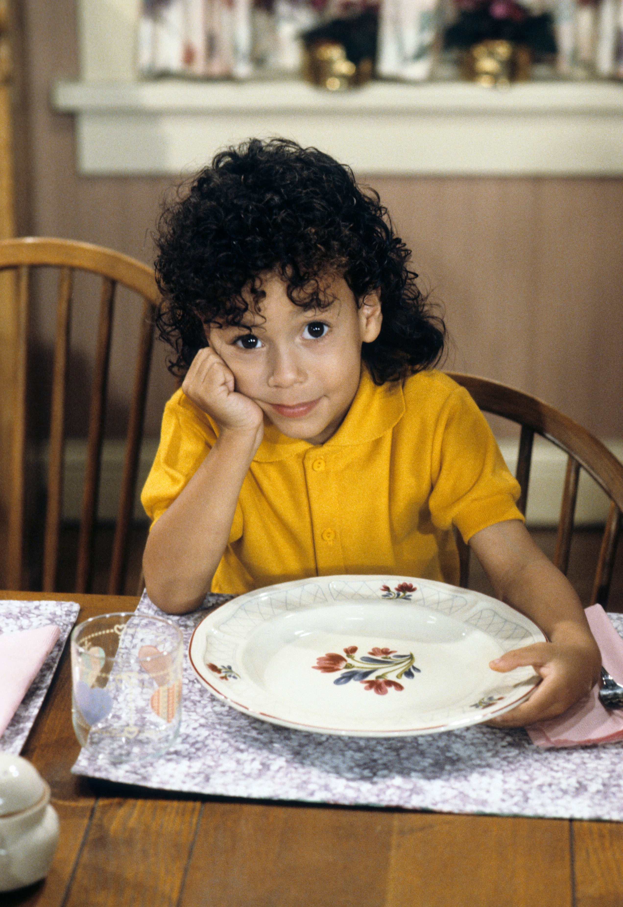 Child with curly hair sits at a dining table holding a floral plate, looking directly at the camera with a curious expression