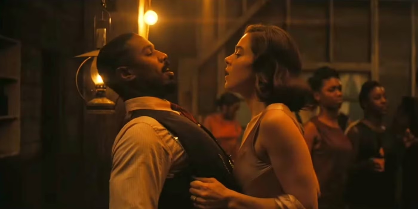 Two actors in an intense scene, one leaning back and the other close, in a dimly lit room with background extras