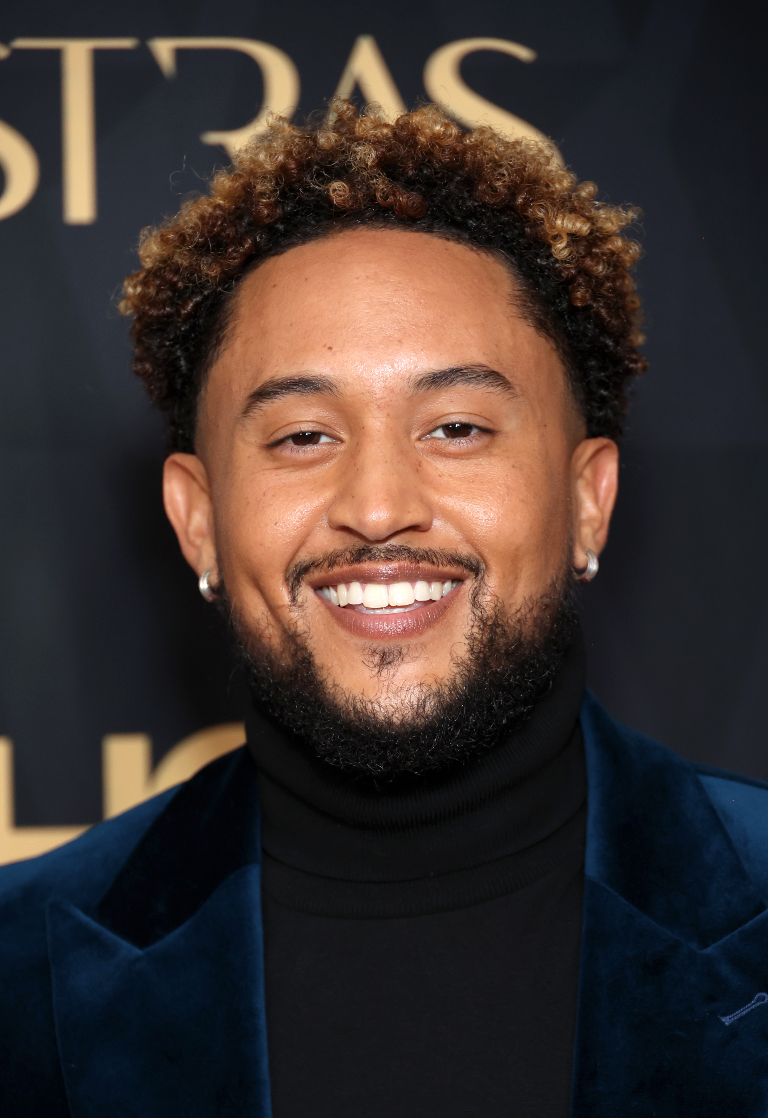 A man with curly hair smiles on the red carpet, wearing a velvet jacket and turtleneck
