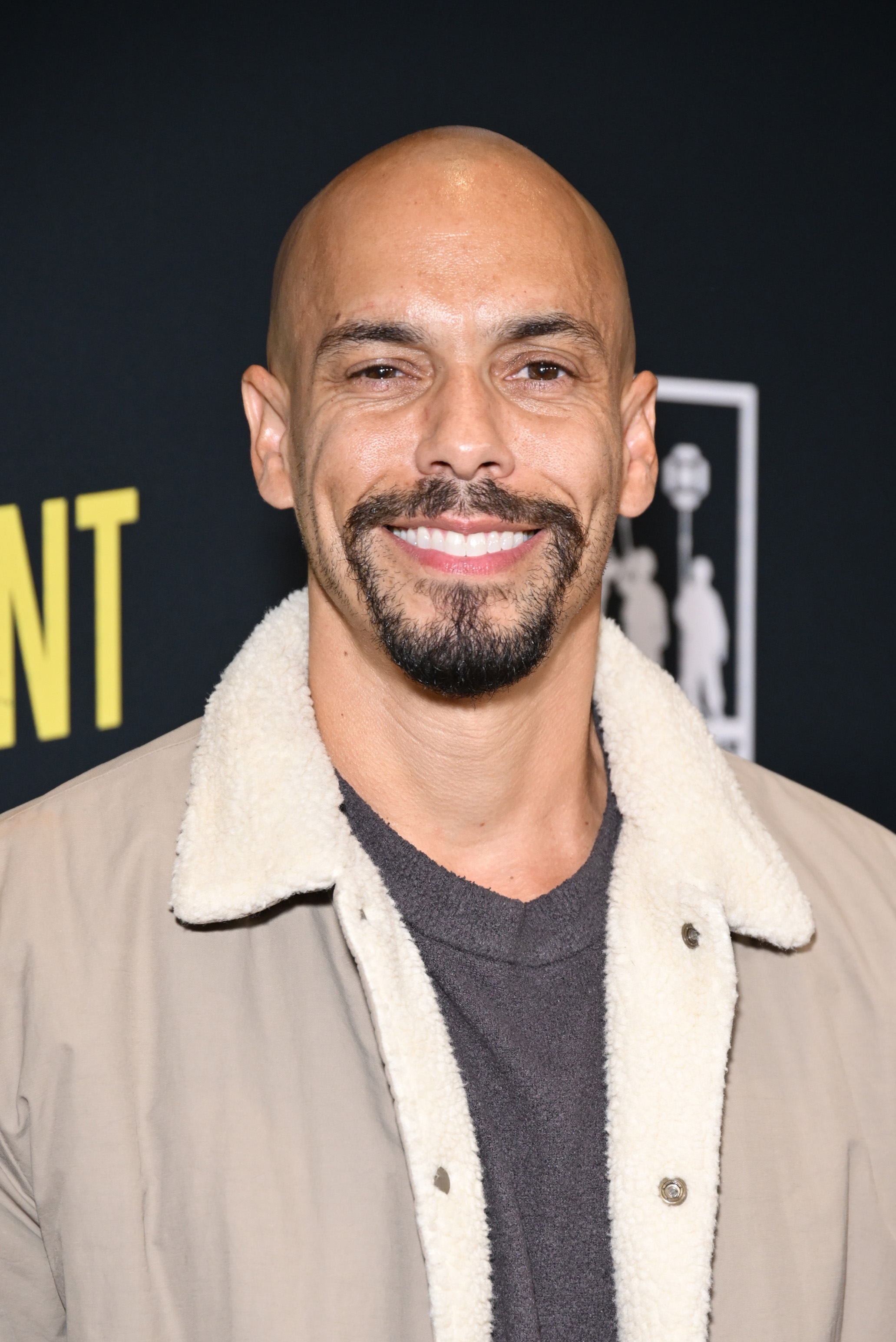 Smiling person with shaved head and beard in a casual coat with a white fleece collar at a celebrity event