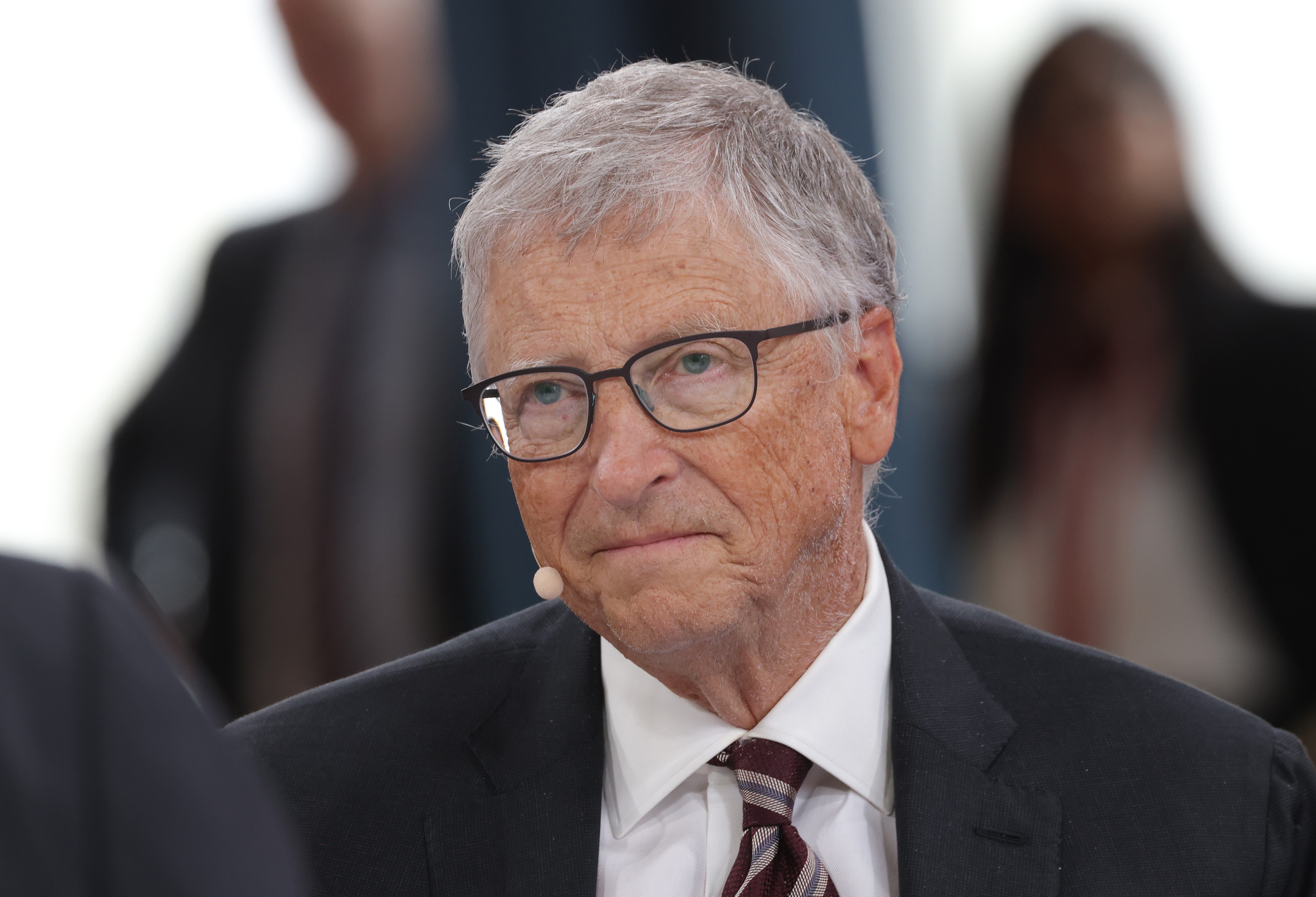 Bill Gates in a suit and tie speaking in a formal setting with a microphone attached to his ear