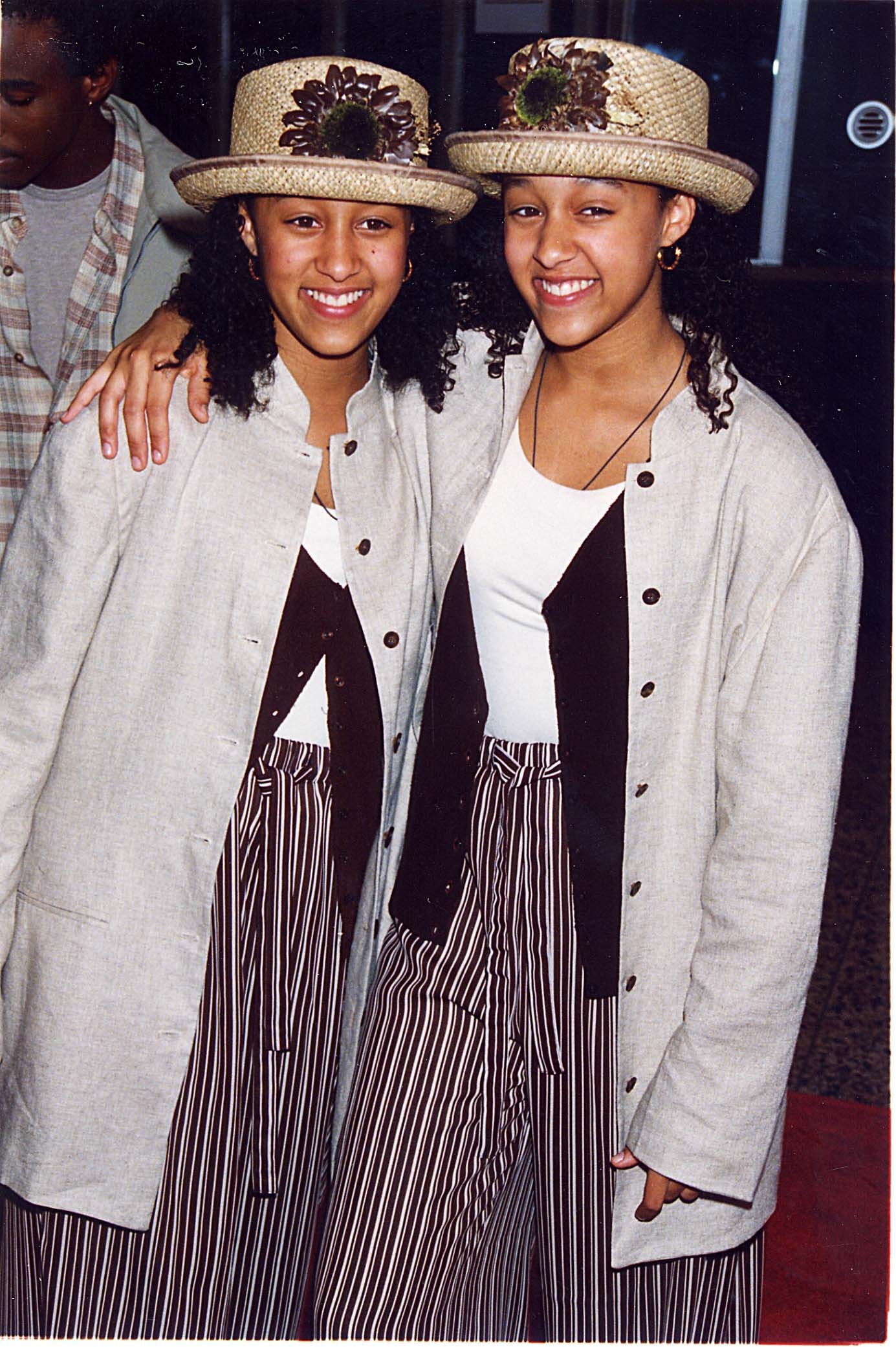 Two individuals on a red carpet wearing matching hats, beige jackets, and striped pants, smiling and posing for a photo