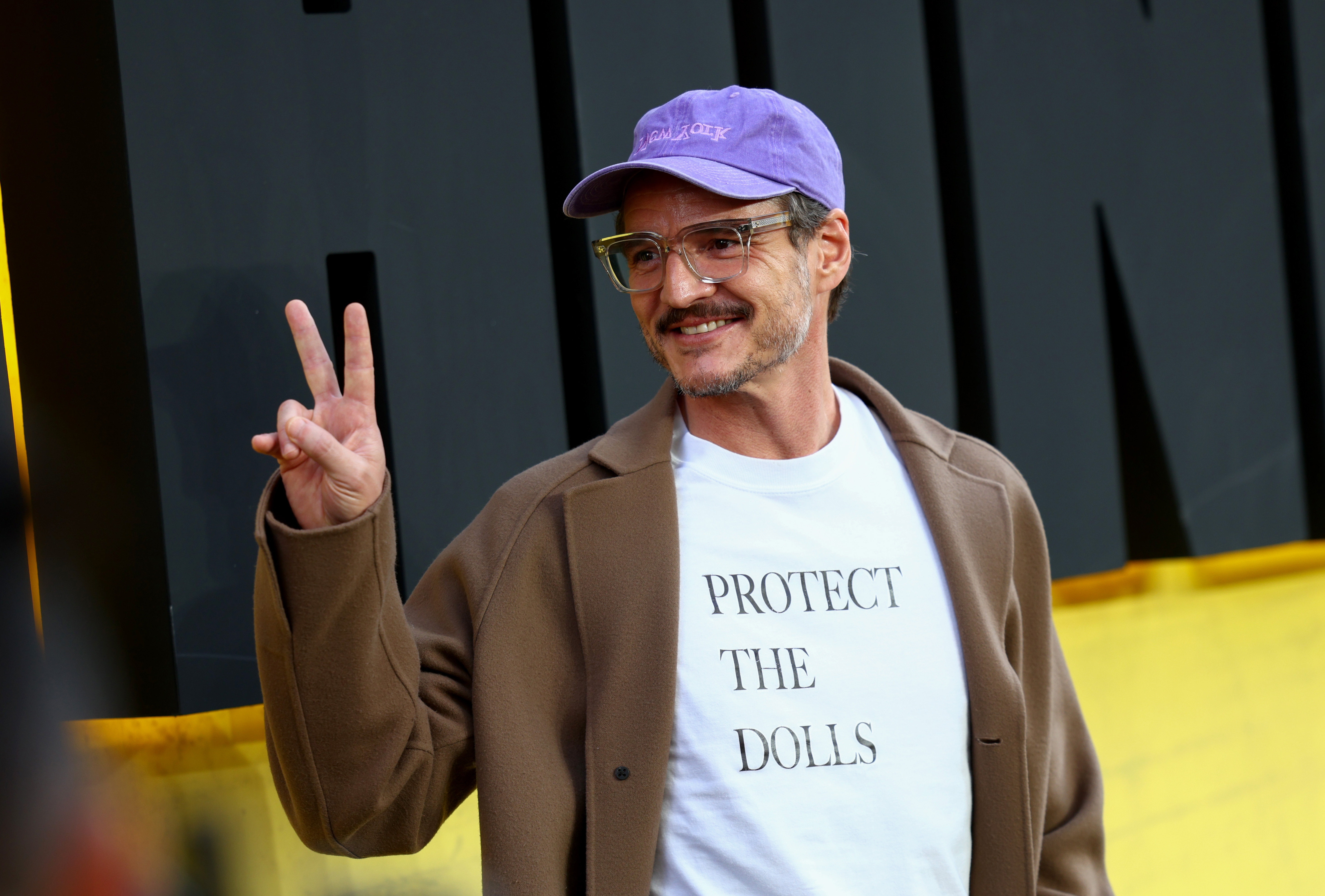 Pedro wearing glasses, a purple cap, and a brown jacket over a "Protect the Dolls" shirt, gestures peace sign