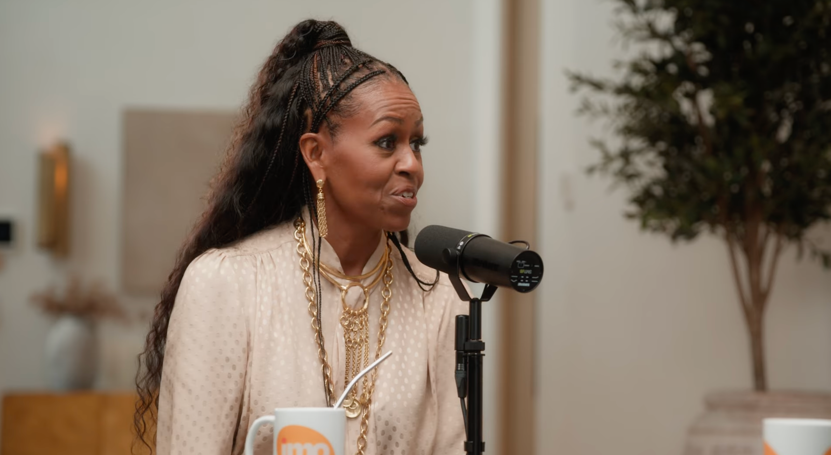 Person with long hair speaks into a microphone during a podcast, wearing a cream blouse and gold jewelry, seated at a table with a mug