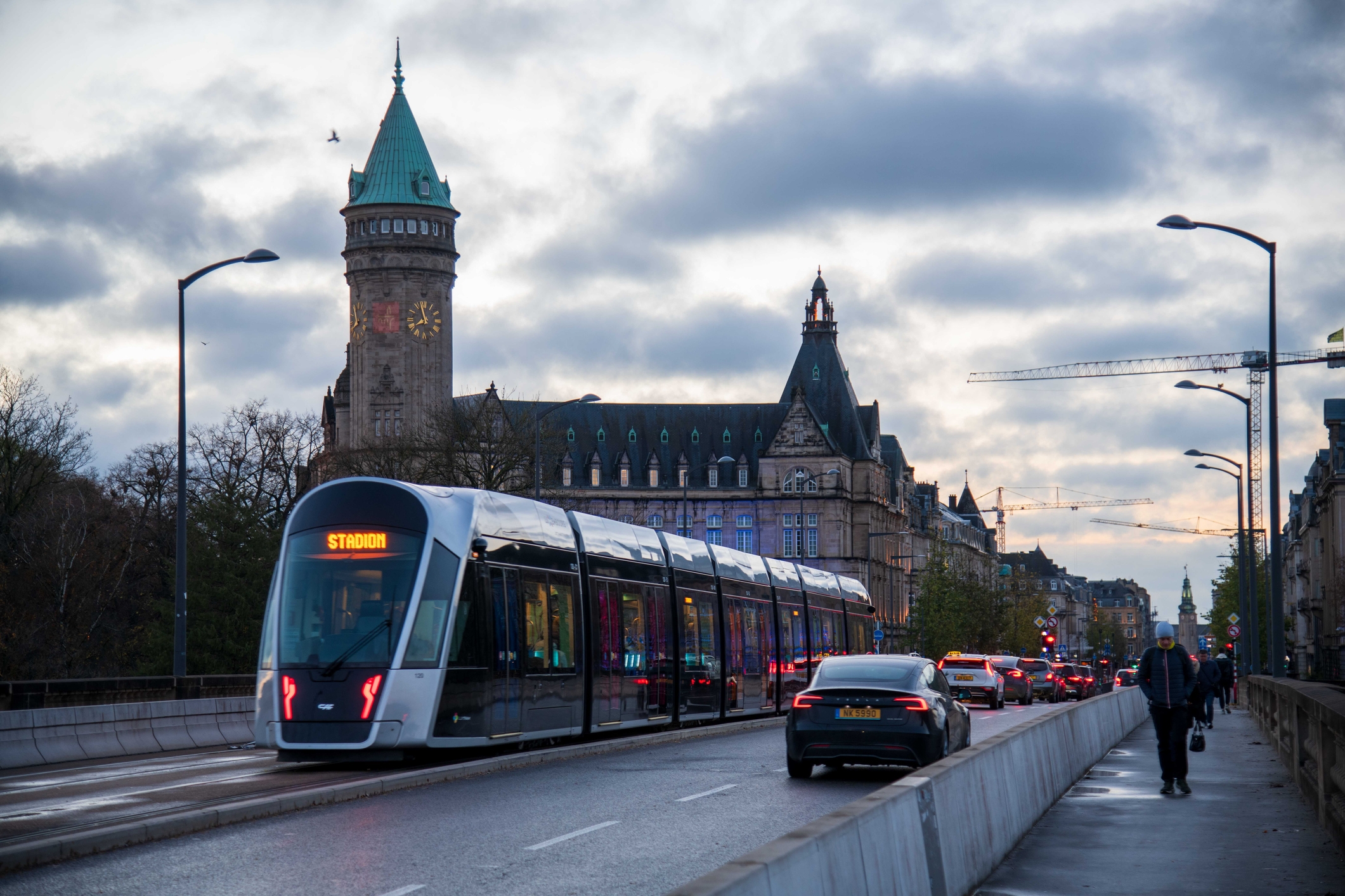 A modern tram travels on a city street beside a historic building with a large clock tower, under a cloudy sky