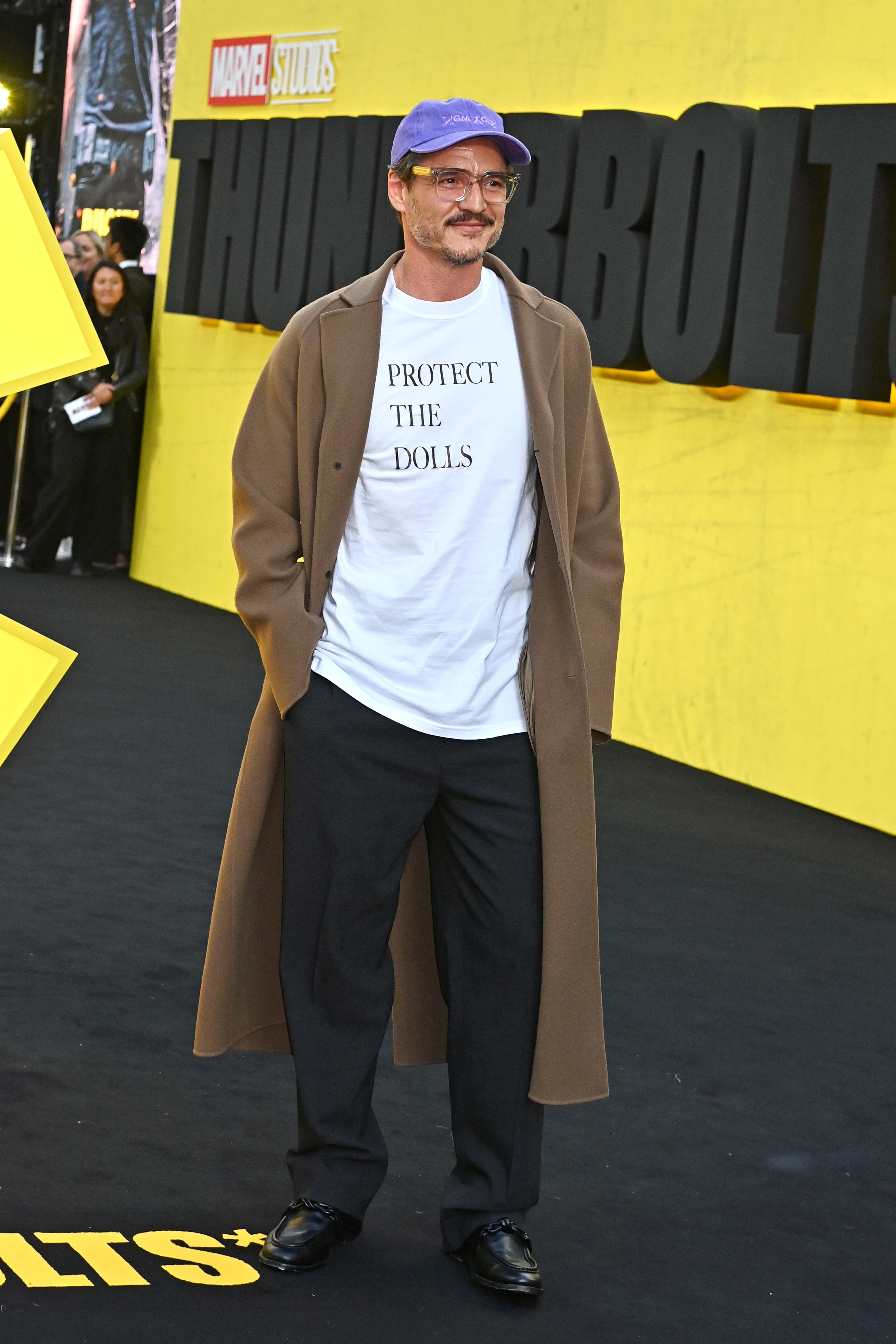 Pedro wearing casual outfit with a camel coat, graphic tee, black pants, and purple cap