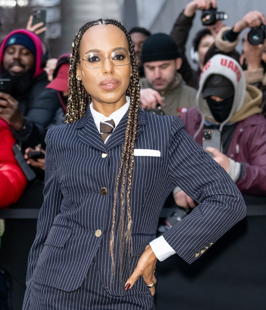 A person in a pinstripe suit, glasses, and long braids poses confidently on a red carpet while photographers capture the moment