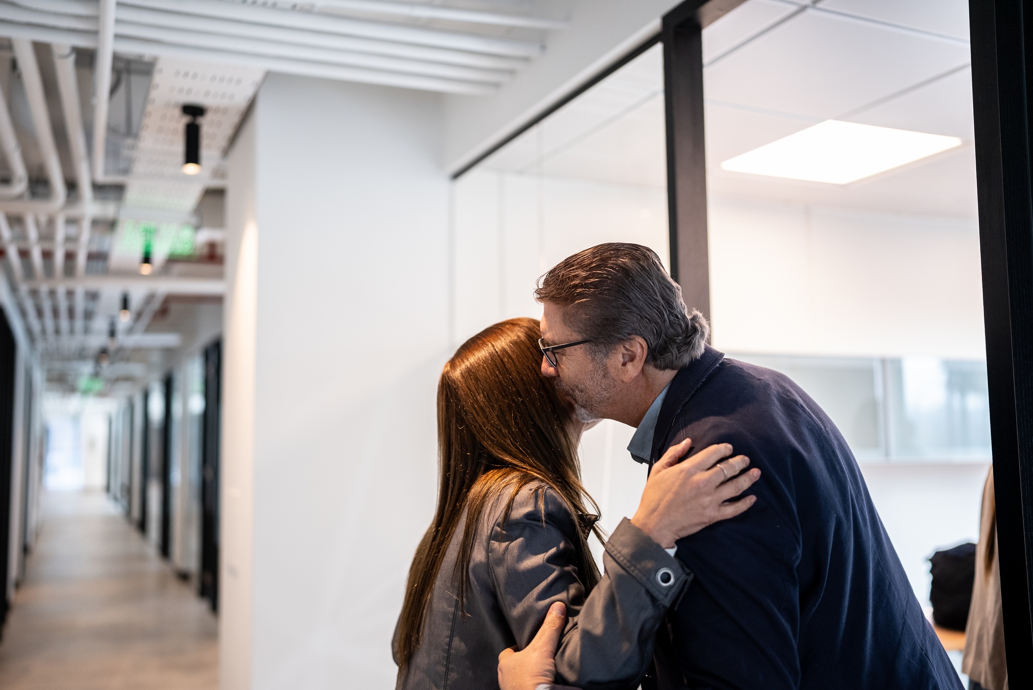 Two people hugging in a professional office hallway, expressing a warm greeting or farewell