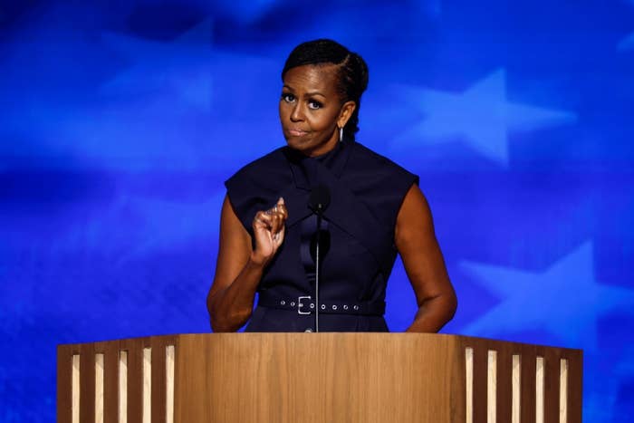 A person speaks at a podium against a backdrop with star patterns, wearing a sleeveless high-collar dress, conveying a confident demeanor