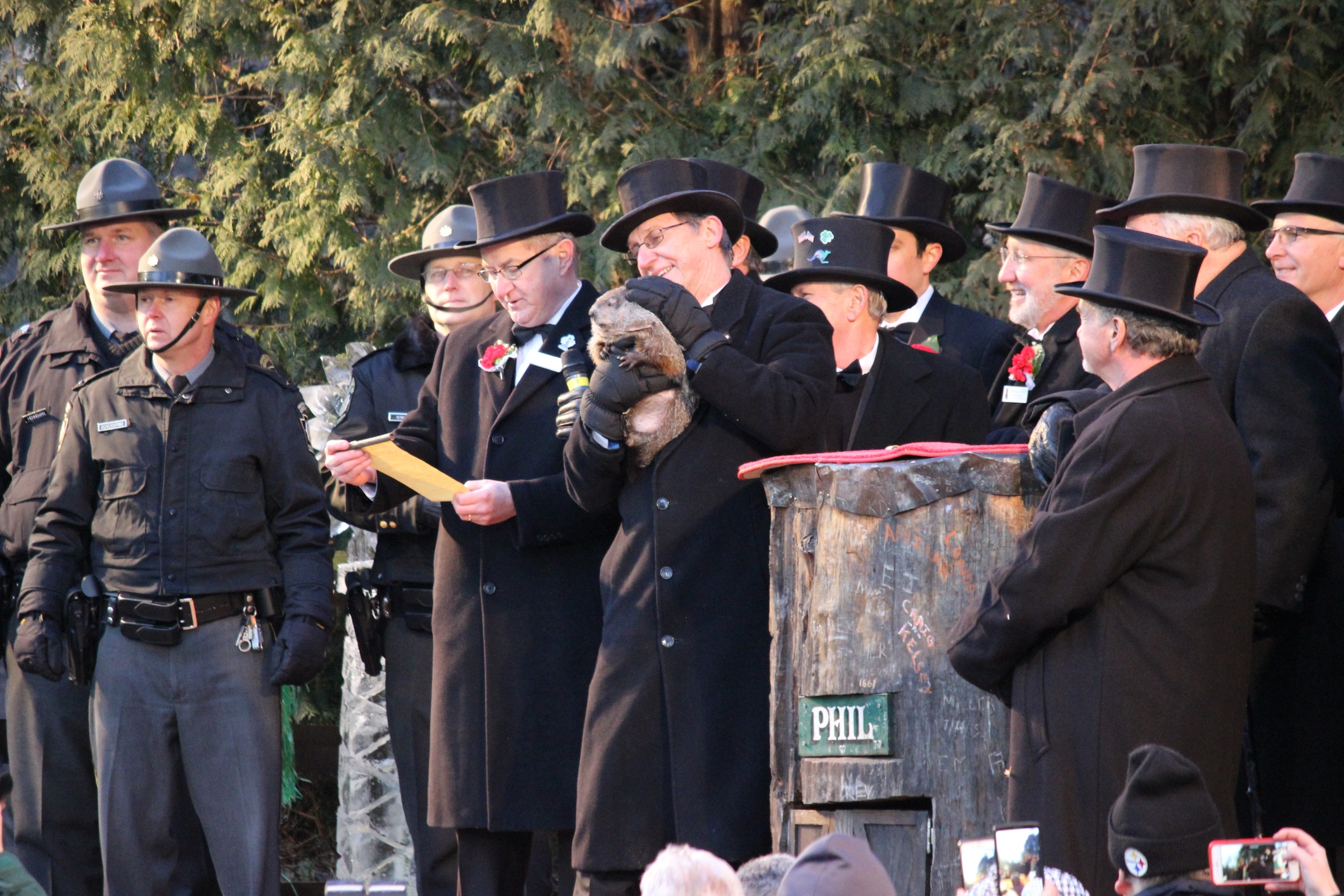 A group of people in formal coats and top hats celebrate Groundhog Day with a groundhog. They stand near a tree stump marked &quot;Phil.&quot;