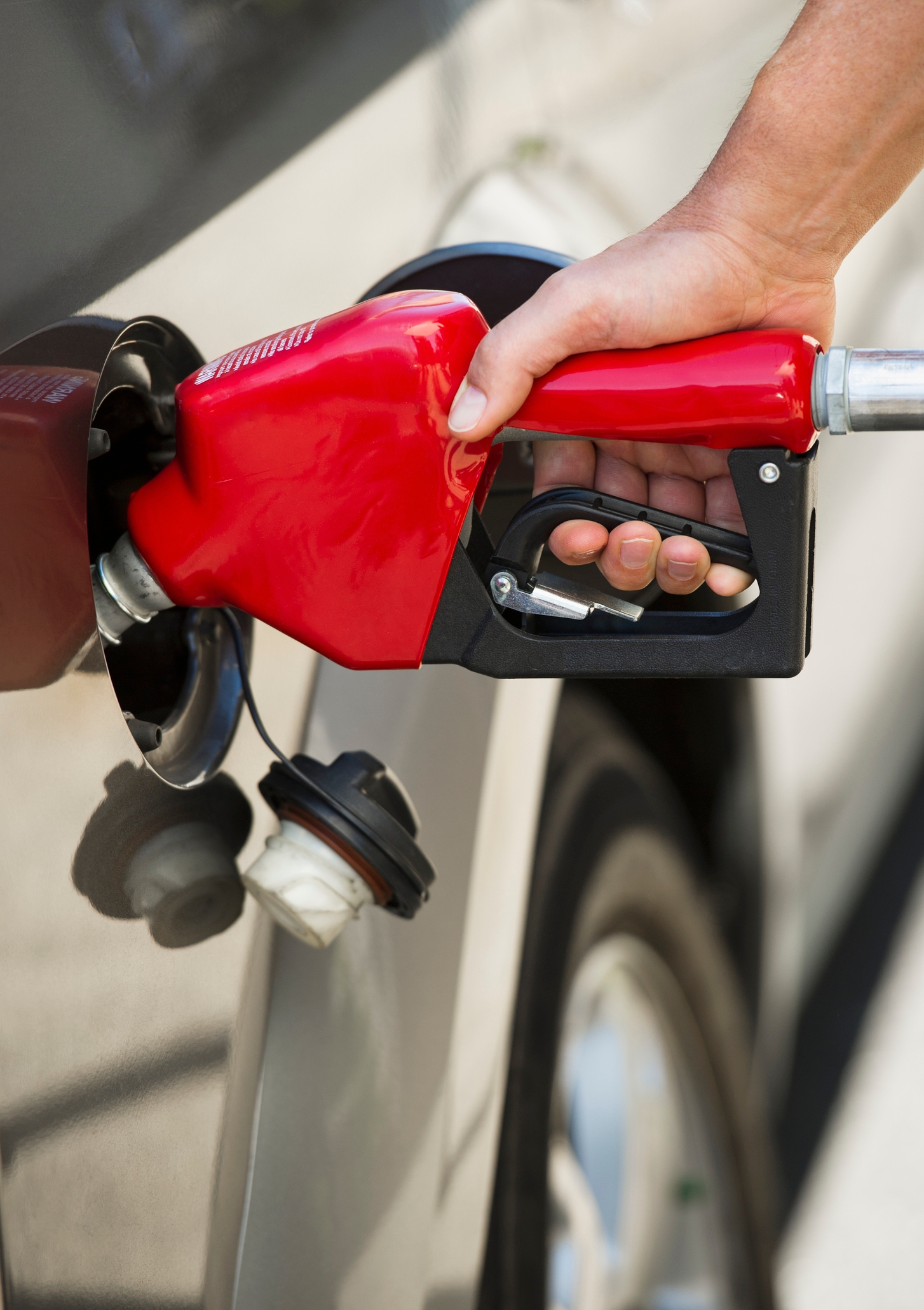 Hand holding a fuel pump nozzle inserting it into a car's gas tank, with the cap hanging nearby, illustrating refueling process