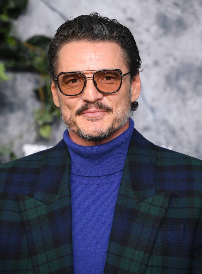 Pedro Pascal wearing large glasses, a blue turtleneck, and a checkered blazer smiles at a media event