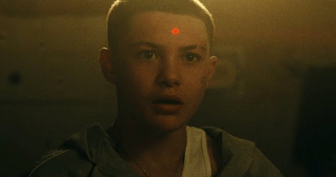Ashtray from "Euphoria" with a buzz cut looks surprised, wearing a casual top in dim lighting, with a red dot on forehead