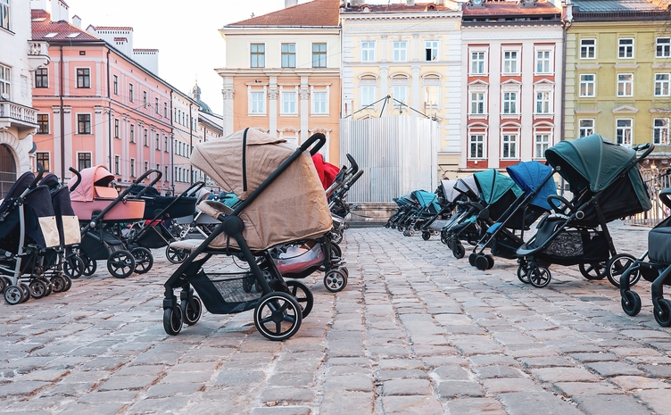A plaza filled with numerous empty baby strollers on a cobblestone street, surrounded by colorful historic buildings
