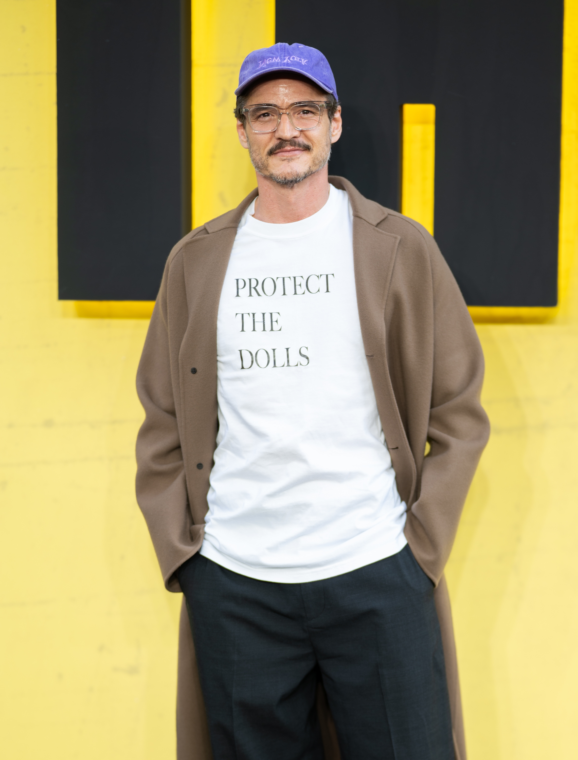 Pedro in a long coat, white t-shirt with "Protect the Dolls" text, and a cap