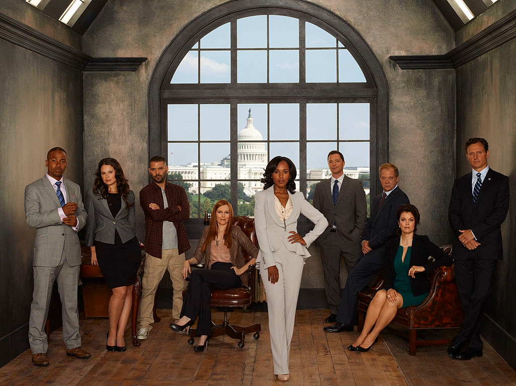 A group of professionally dressed individuals pose in an indoor setting with a window view of the U.S. Capitol in the background