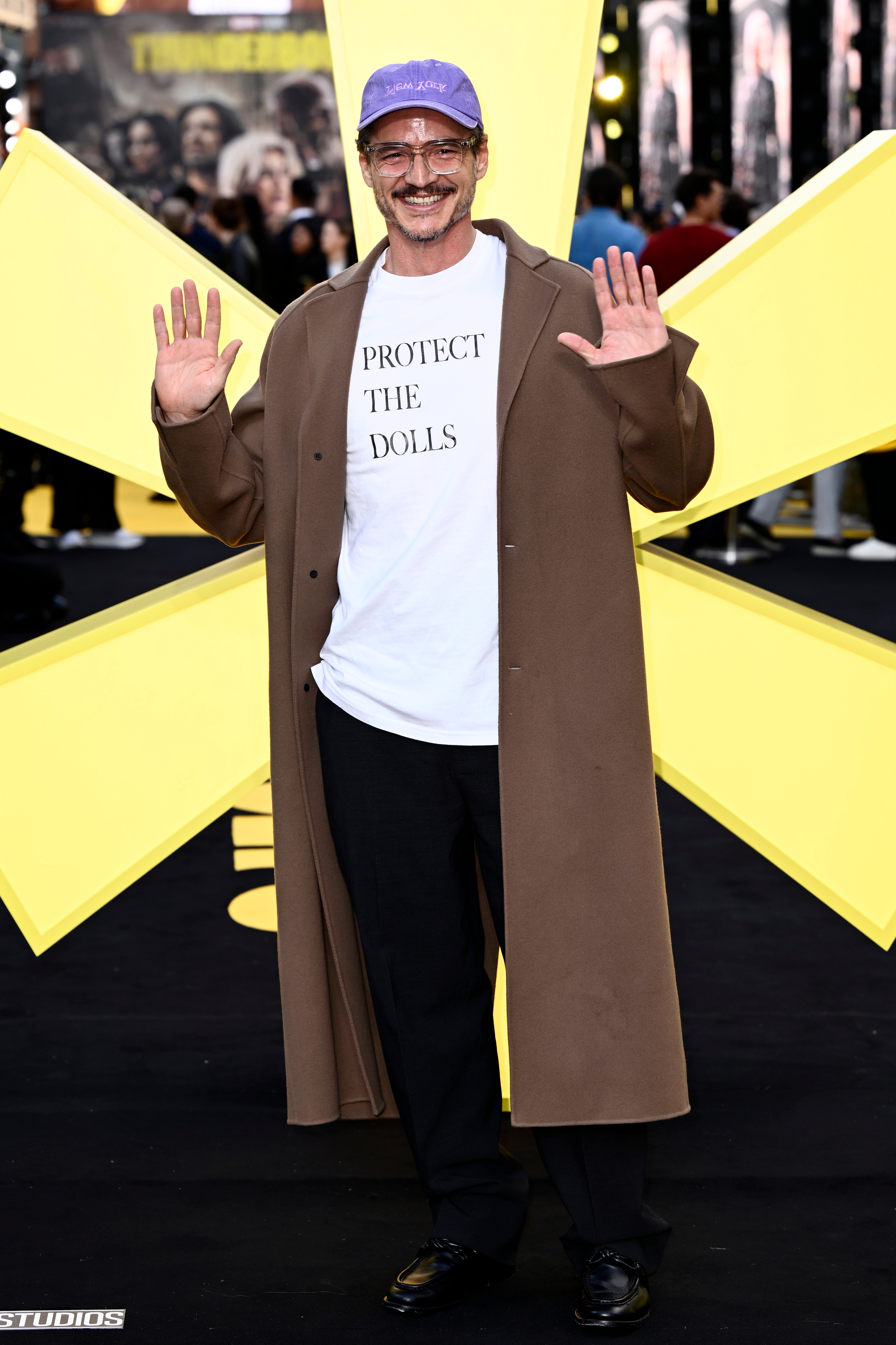 Pedro in a long coat, graphic t-shirt, and cap smiling and waving on a black carpet event