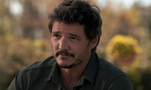 A person with facial hair, wearing a collared shirt, is outside, looking thoughtfully ahead. Trees and blurred foliage are in the background