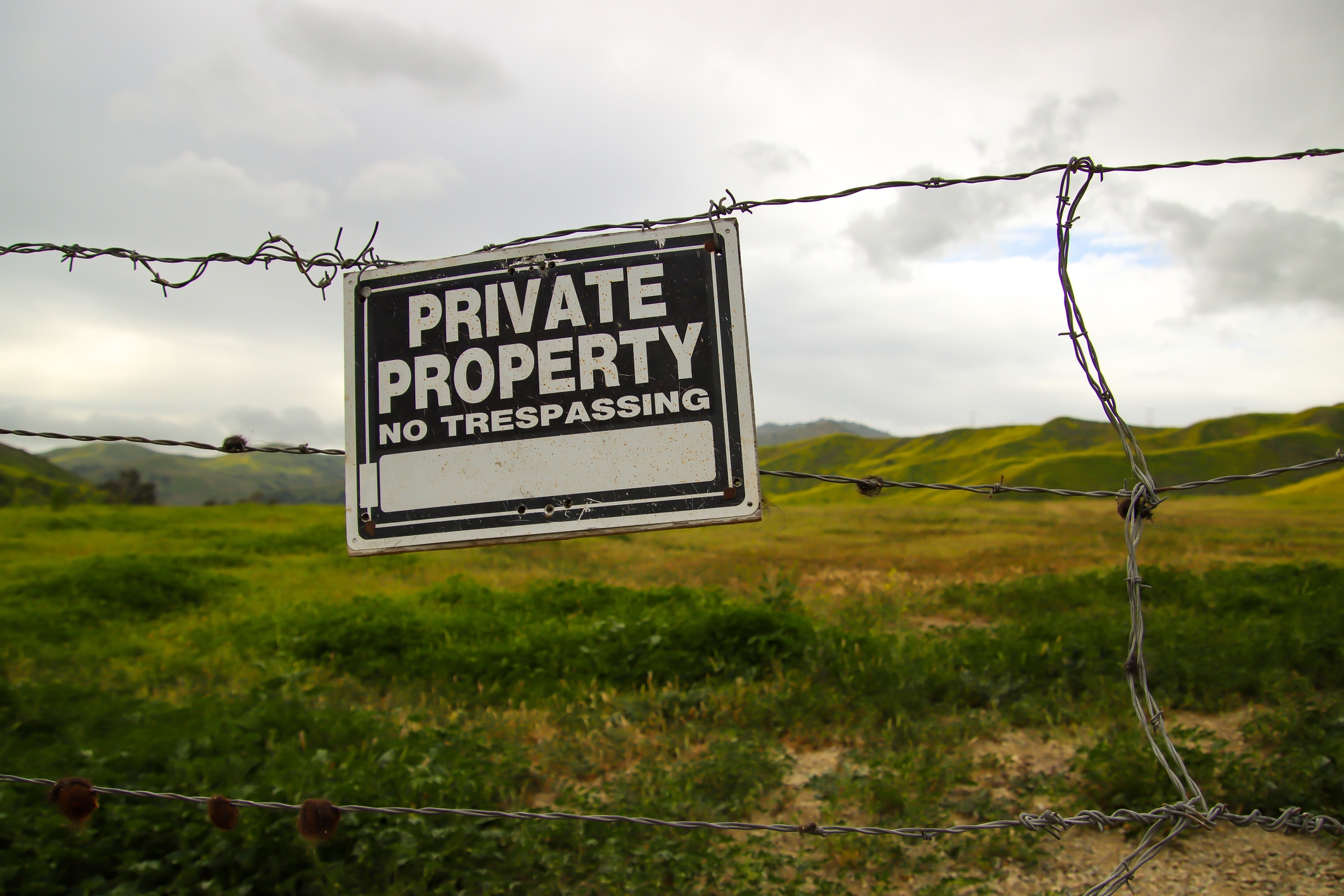 Sign on a barbed wire fence reads, &quot;Private Property, No Trespassing,&quot; with a grassy landscape and hills in the background