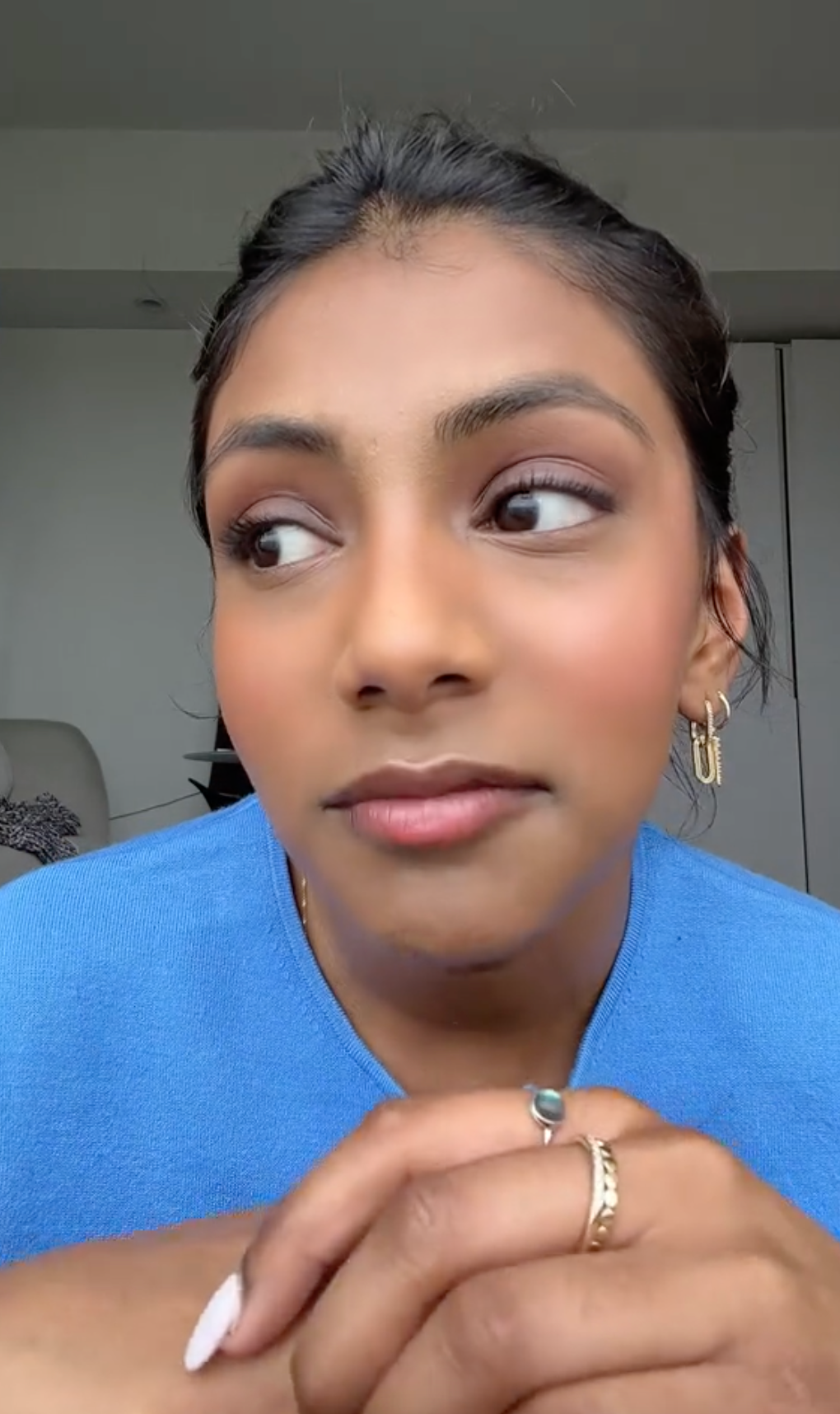 Charithra Chandran with stud earrings and a ring, looking to the side, wearing a casual top, sitting indoors