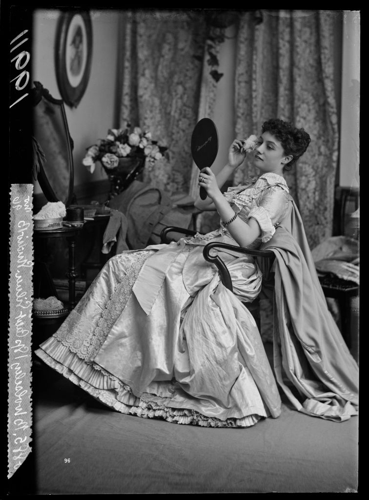 A woman in elegant, vintage attire sits in an armchair, holding a hand mirror, surrounded by draped fabric and floral decor