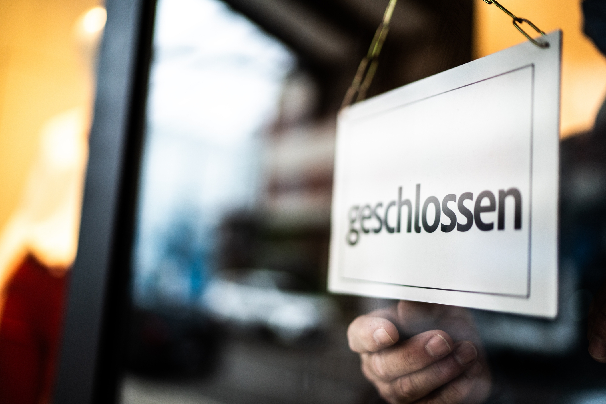A hand holds a &quot;geschlossen&quot; sign in a shop window, indicating the store is closed