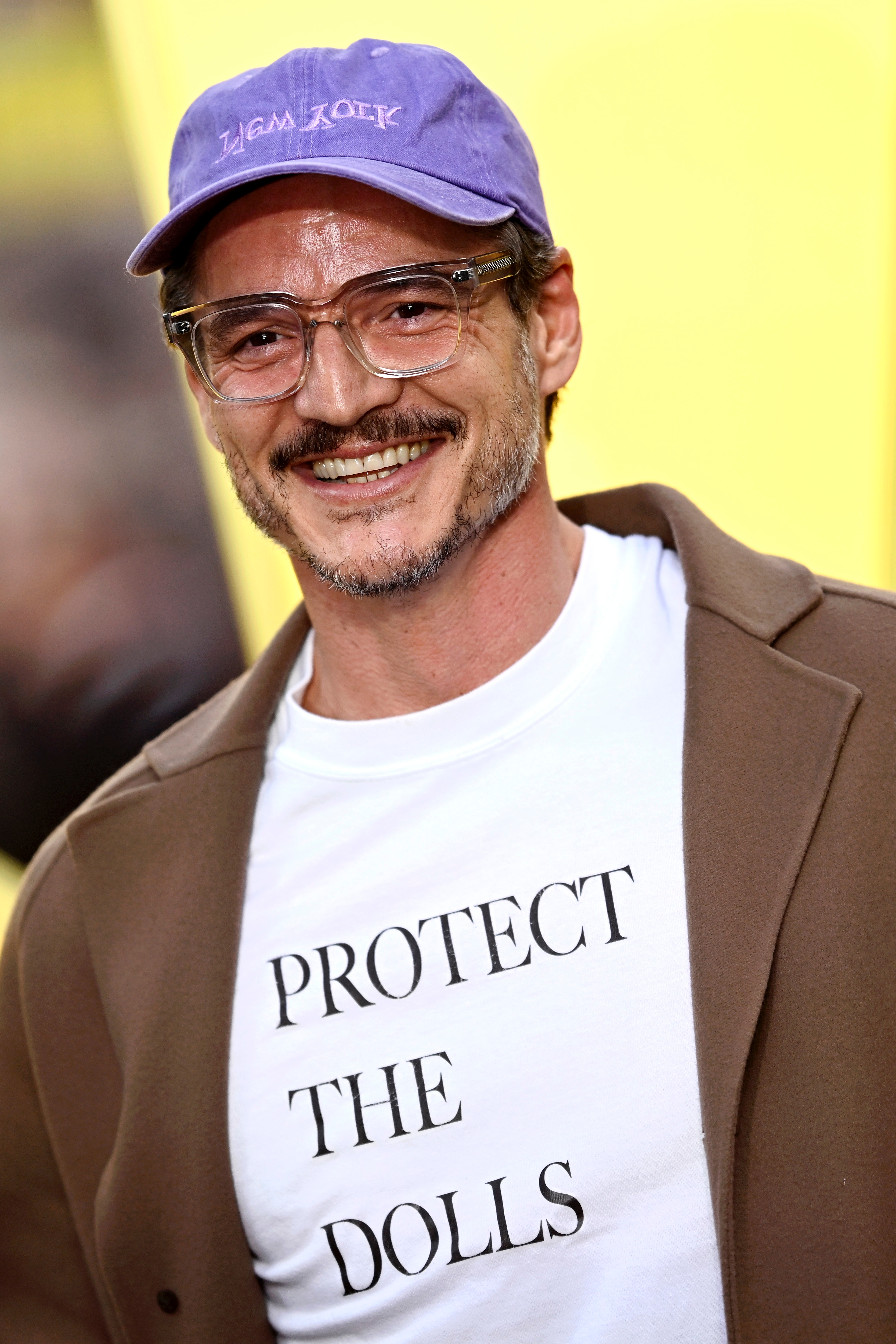 Pedro smiling, wearing glasses, a cap, and a white shirt with "Protect the Dolls" text, layered under a brown coat