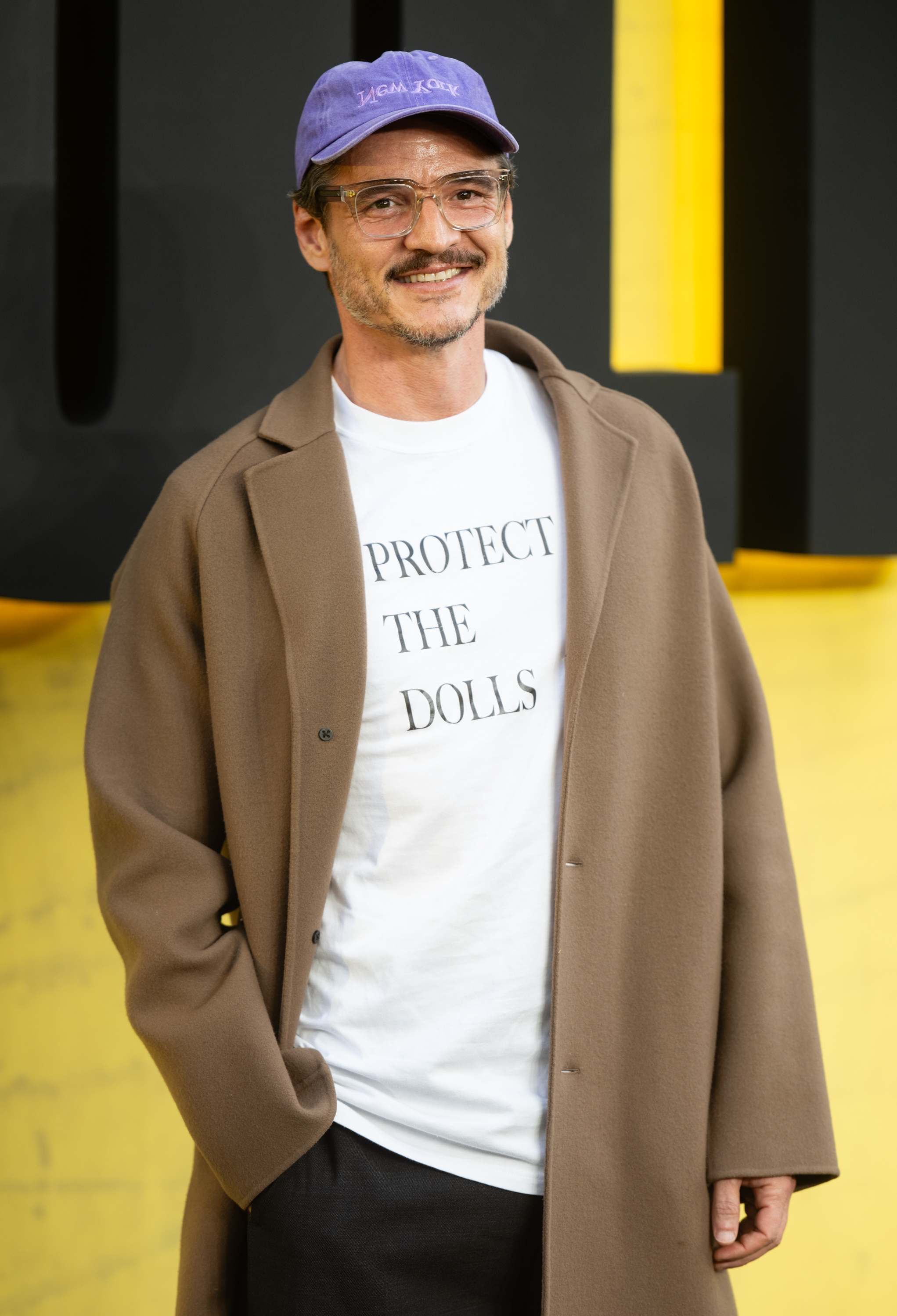 Pedro in a casual outfit with a brown coat, glasses, and a purple cap smiles while wearing a t-shirt that reads "Protect the Dolls."