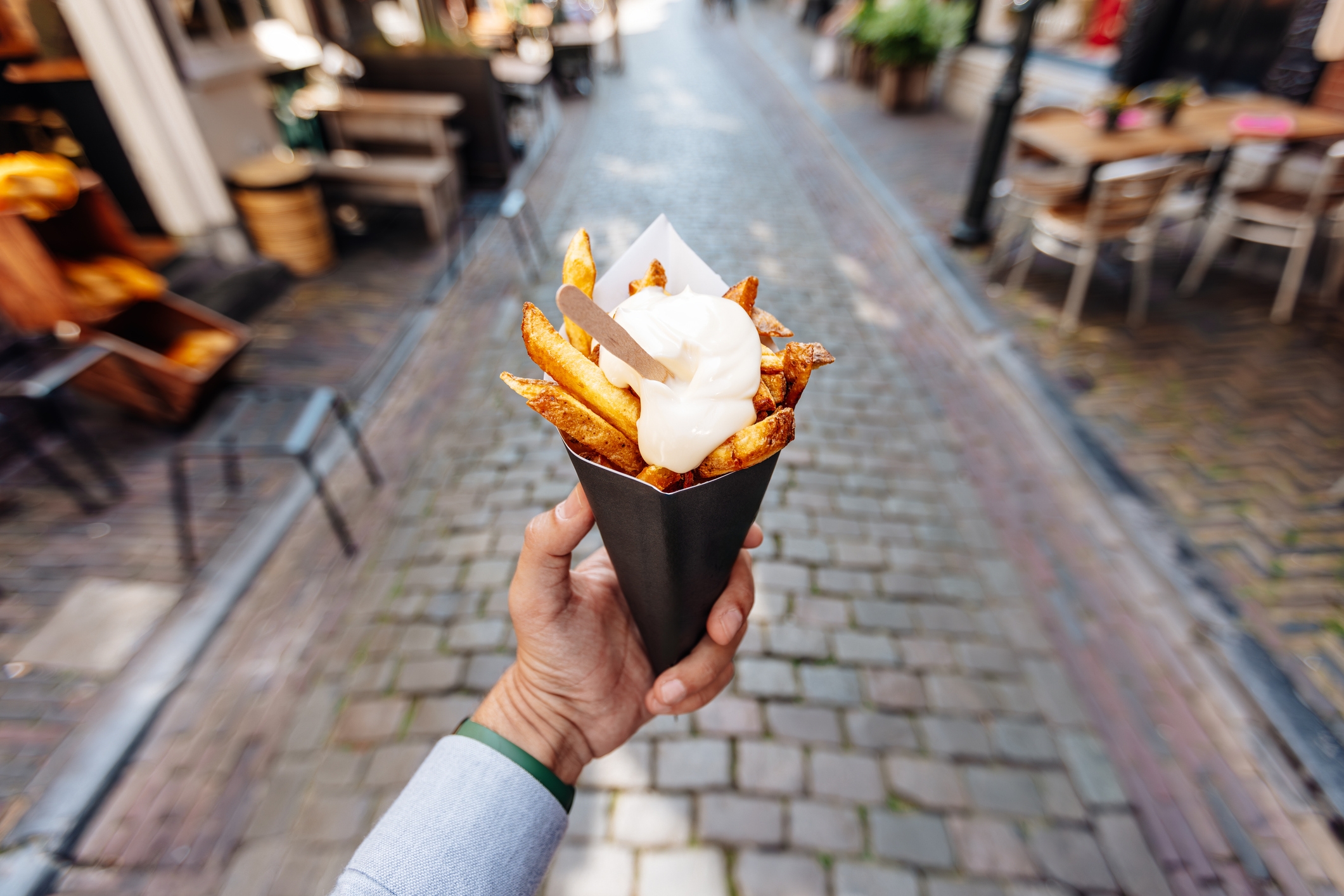 Hand holding cone of fries topped with mayonnaise on a cobblestone street